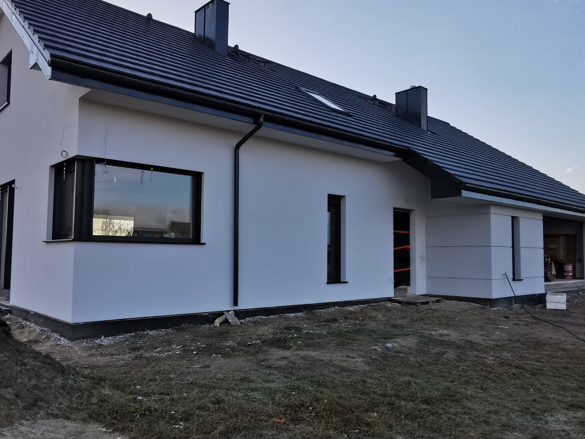 Nowoczesny dom jednorodzinny z białą elewacją, czarną stolarką okienną i dachem. Widoczne detale wykończeniowe i otoczenie budynku w trakcie prac.