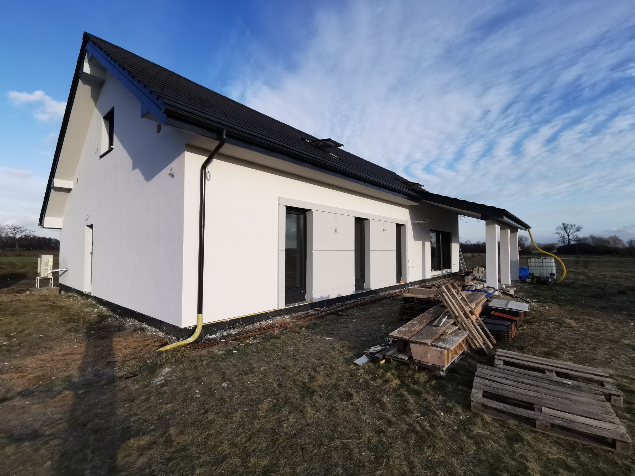 Nowoczesny dom jednorodzinny z białą elewacją i czarnym dachem, widoczny z boku, z elementami wykończeniowymi i materiałami budowlanymi wokół.