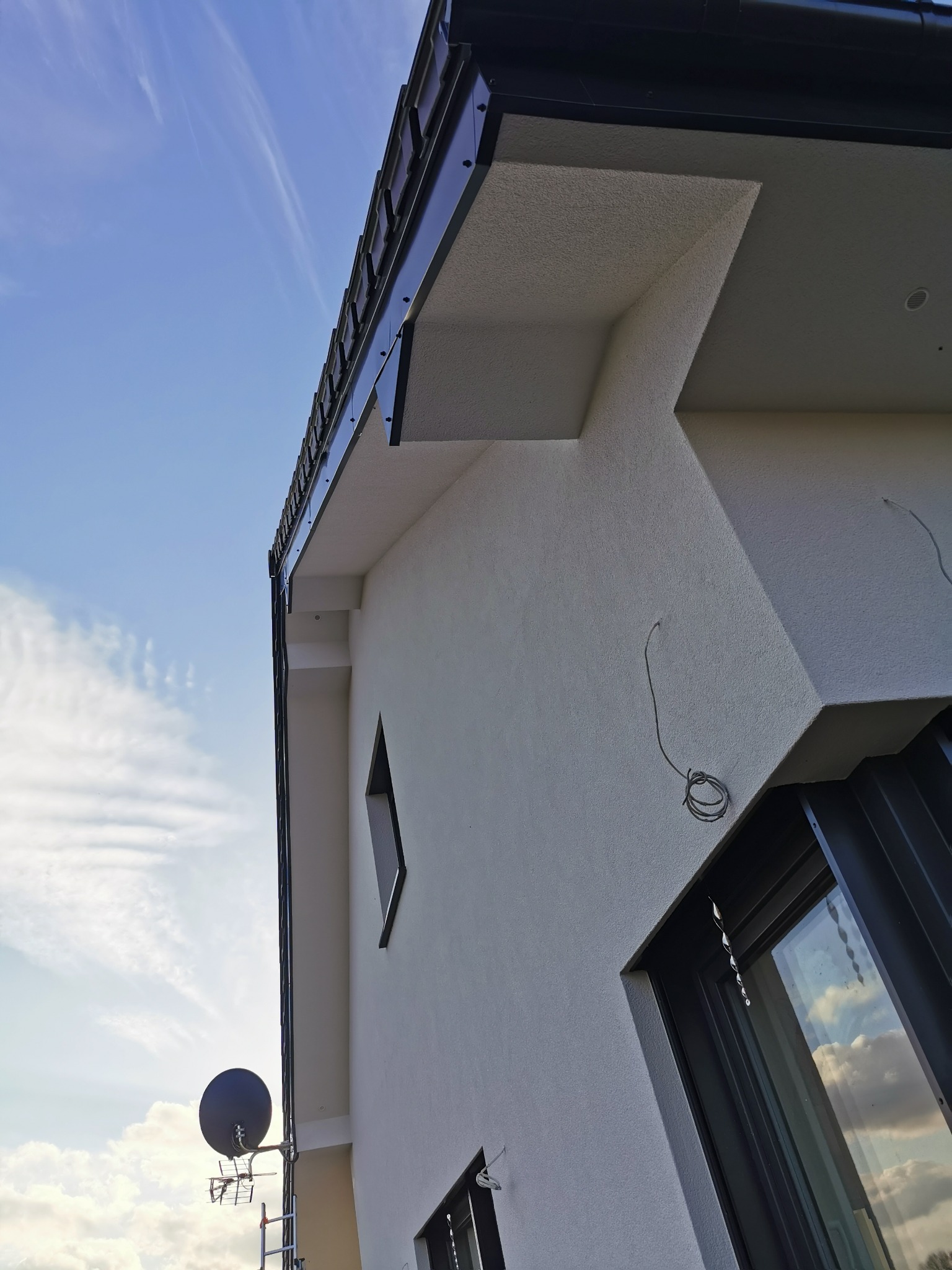 Biała elewacja domu jednorodzinnego z czarnymi elementami wykończeniowymi, widok z dołu na tle błękitnego nieba z chmurami. Widoczne okna i antena satelitarna.