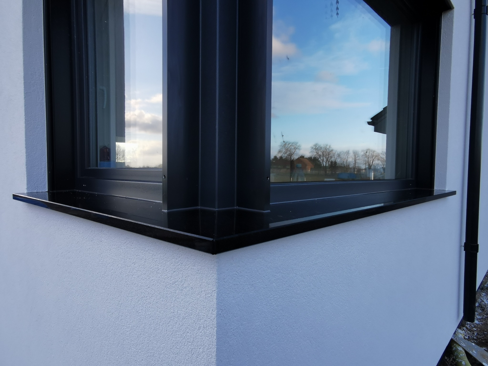 Detal narożnika okna z czarną ramą i parapetem na białej, teksturowanej elewacji. W szybie odbija się błękitne niebo i drzewa.