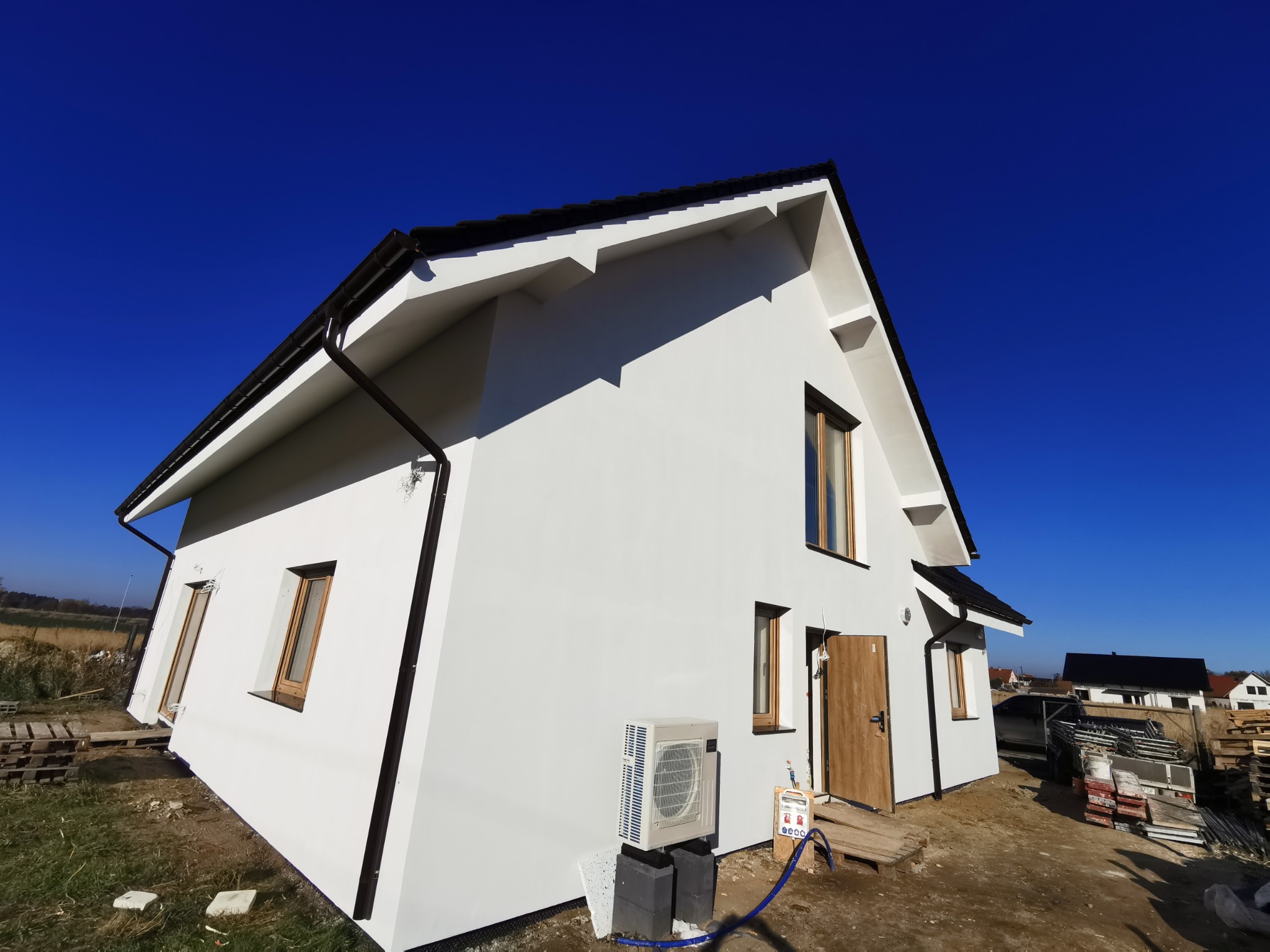 Biały dom jednorodzinny z ciemnym dachem i brązowymi oknami, widziany z niskiej perspektywy, na tle intensywnego błękitu nieba. Widoczny klimatyzator przy ścianie.