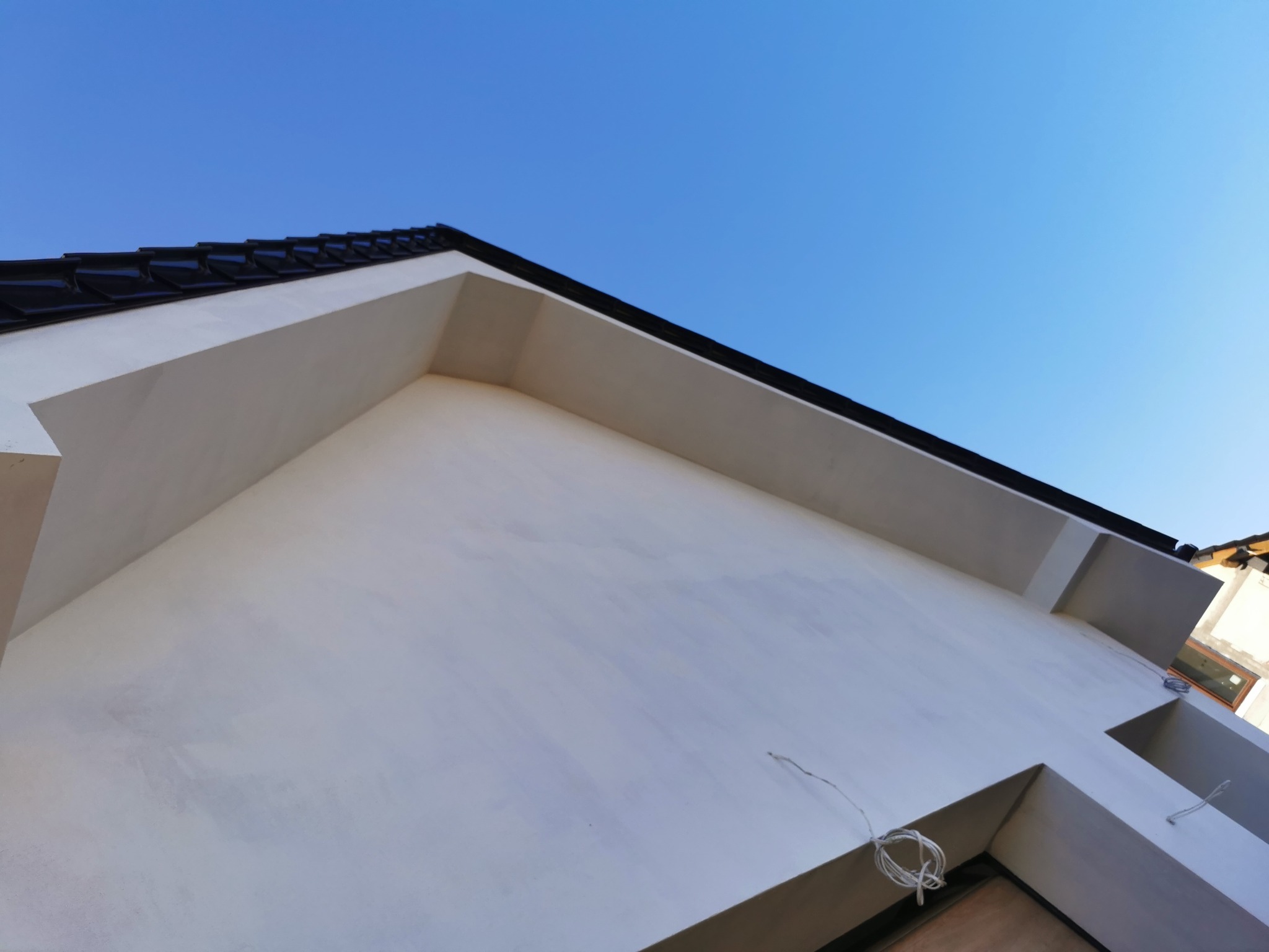 Ściana budynku po tynkowaniu, widok z dołu, z fragmentem dachu i niebieskim niebem w tle. Widoczne przewody elektryczne wystające ze ściany.