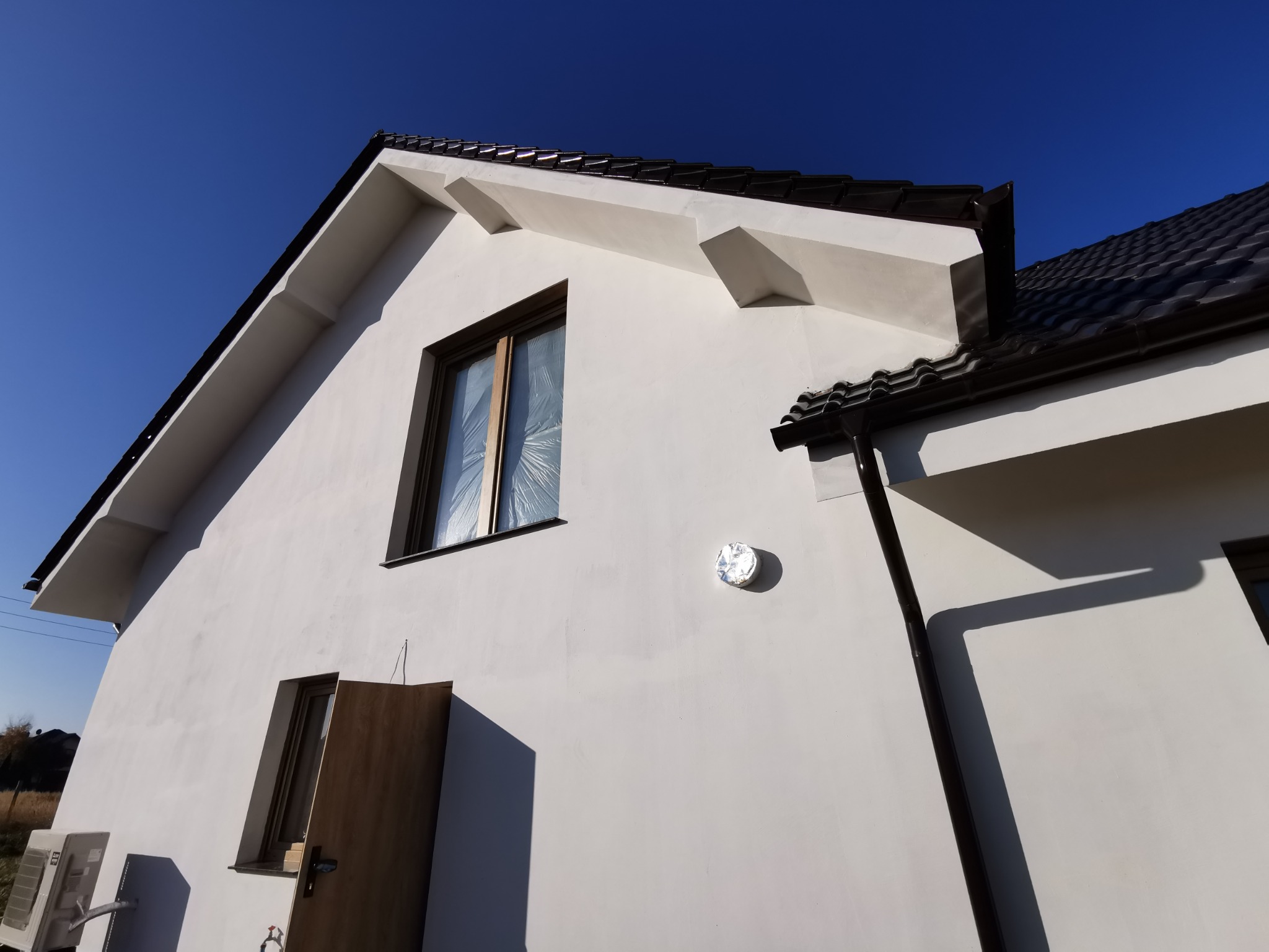 Biała elewacja domu jednorodzinnego z brązową stolarką okienną i dachem z blachodachówki. Okno zabezpieczone folią. Widok z dołu na fragment budynku w słoneczny dzień.
