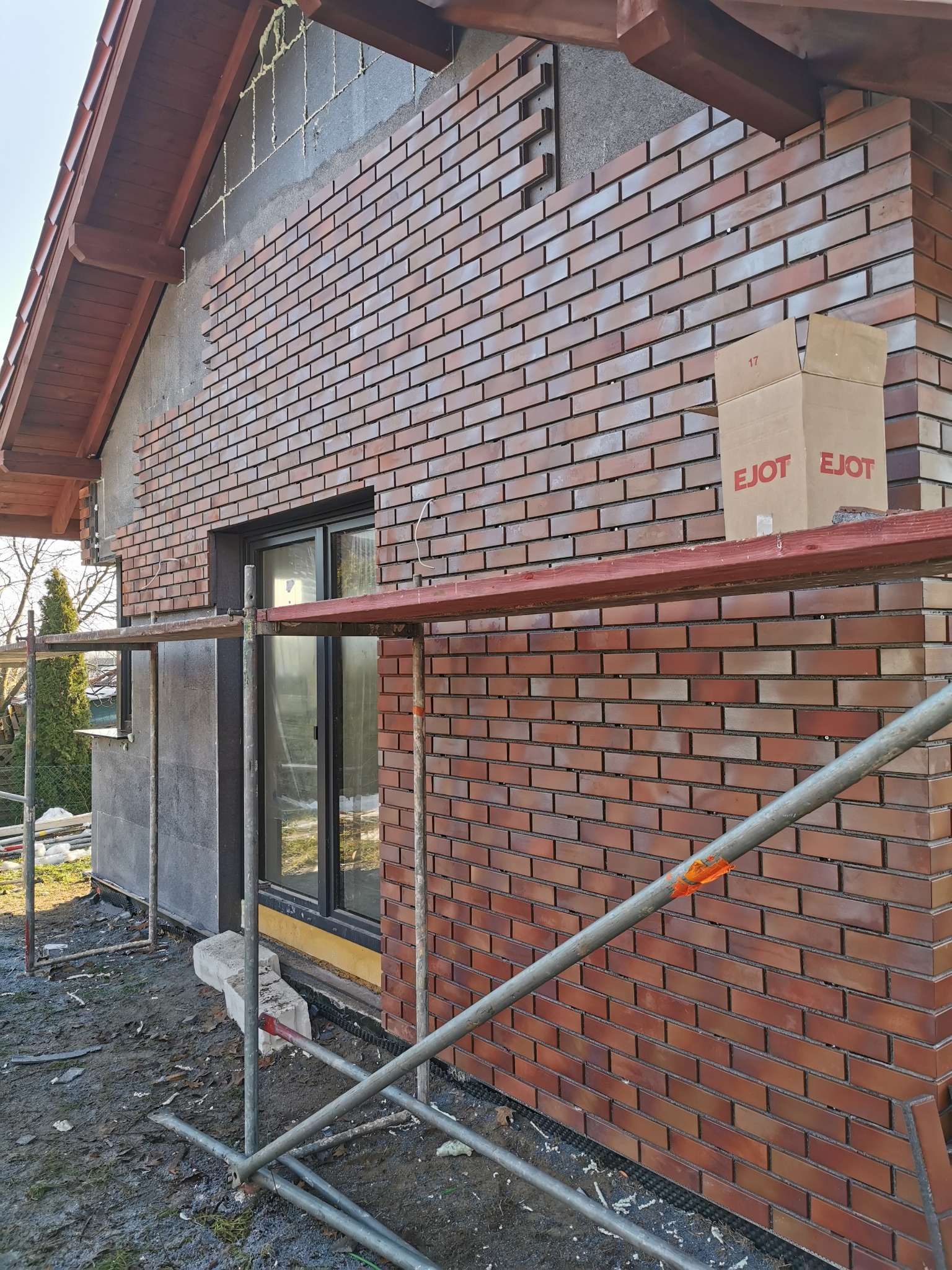 Montaż elewacji z cegły klinkierowej na budynku jednorodzinnym. Widoczne rusztowanie, kartony z materiałem i fragmenty nieocieplonej ściany.