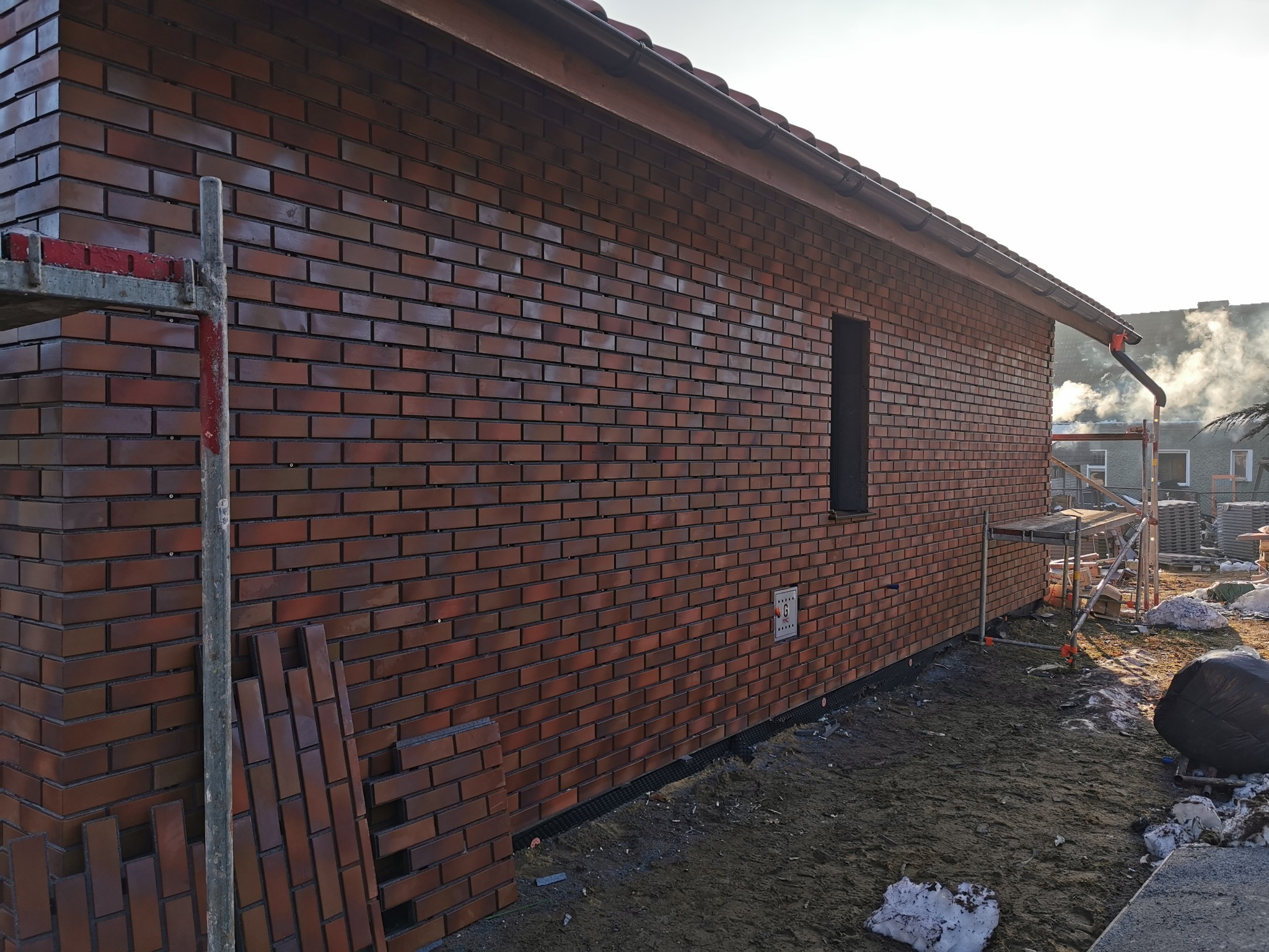 Ściana budynku wykończona czerwoną cegłą klinkierową, widoczne rusztowanie i zapas cegieł. Detal elewacji z wąskim oknem i fragmentem dachu. Słoneczny dzień.