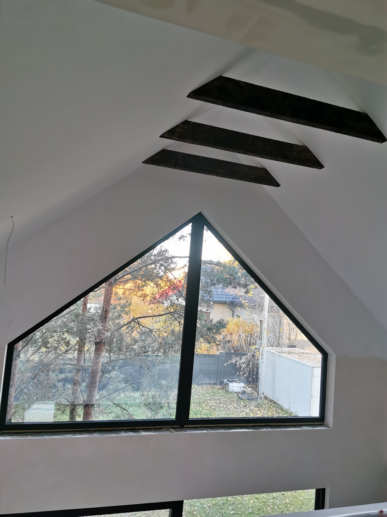 Poddasze z trójkątnym oknem i widokiem na jesienny ogród. Widoczne drewniane belki stropowe i świeżo wykończone ściany.