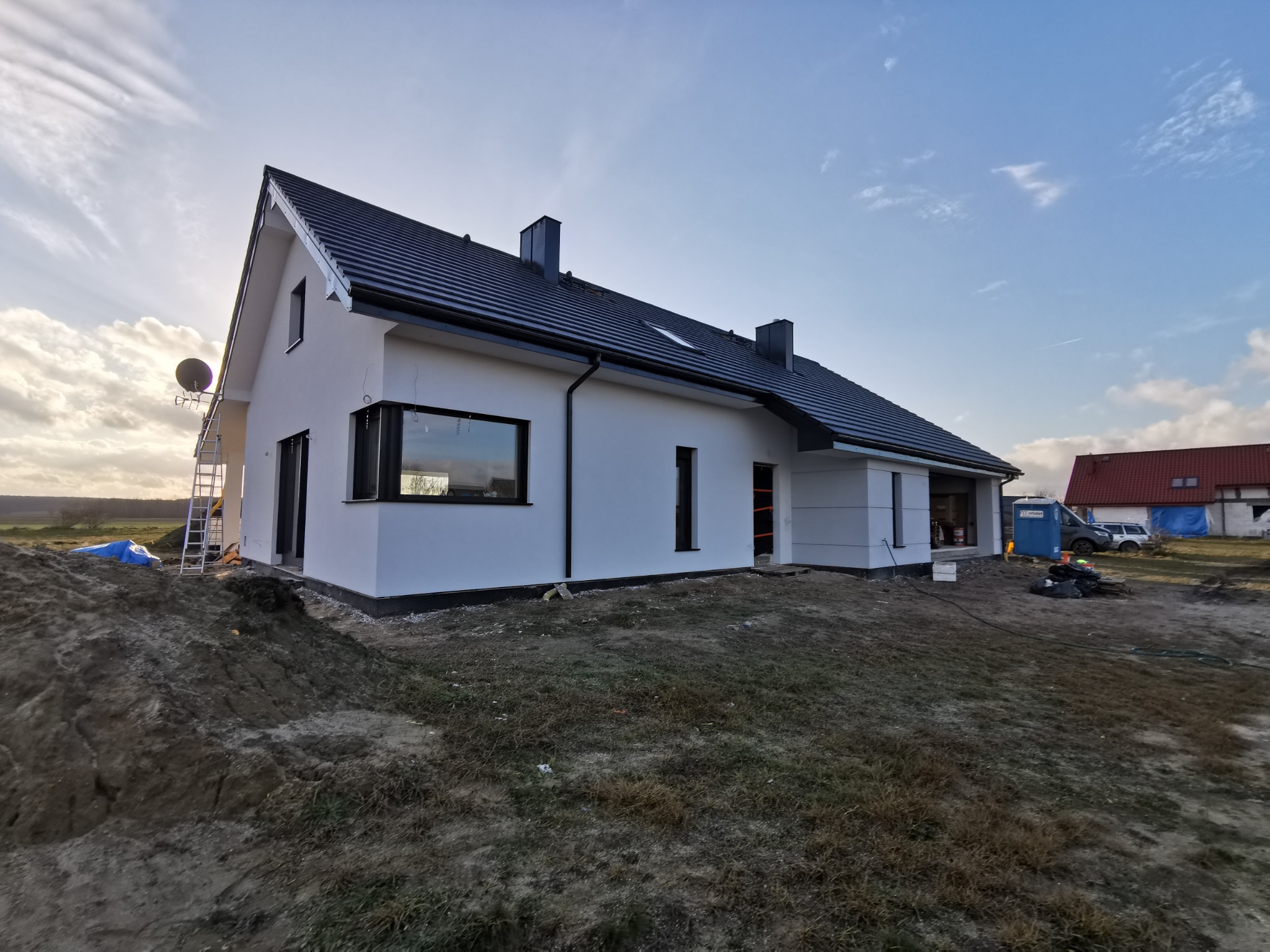Nowoczesny dom jednorodzinny z białymi ścianami i czarnym dachem, widoczny z perspektywy ogrodu. Na pierwszym planie ziemia, w tle błękitne niebo o zmierzchu.