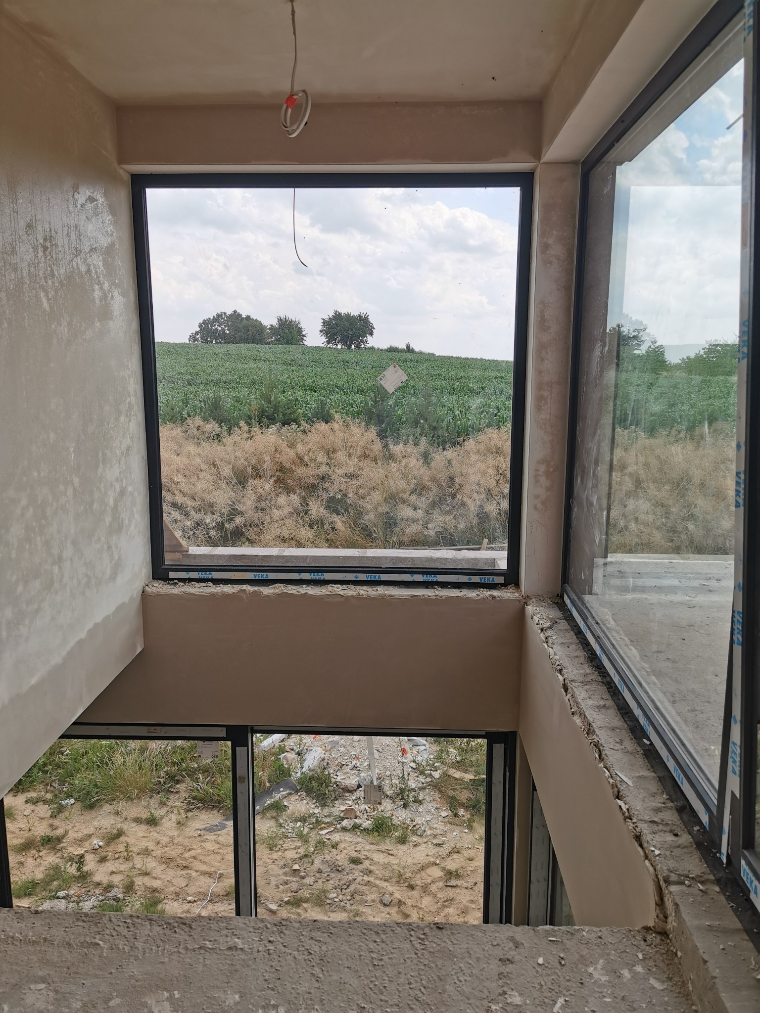 Świeżo otynkowana ściana wewnętrzna, widok na pole uprawne przez czarne ramy okienne. Widoczny surowy stan wykończenia i wystające przewody elektryczne.