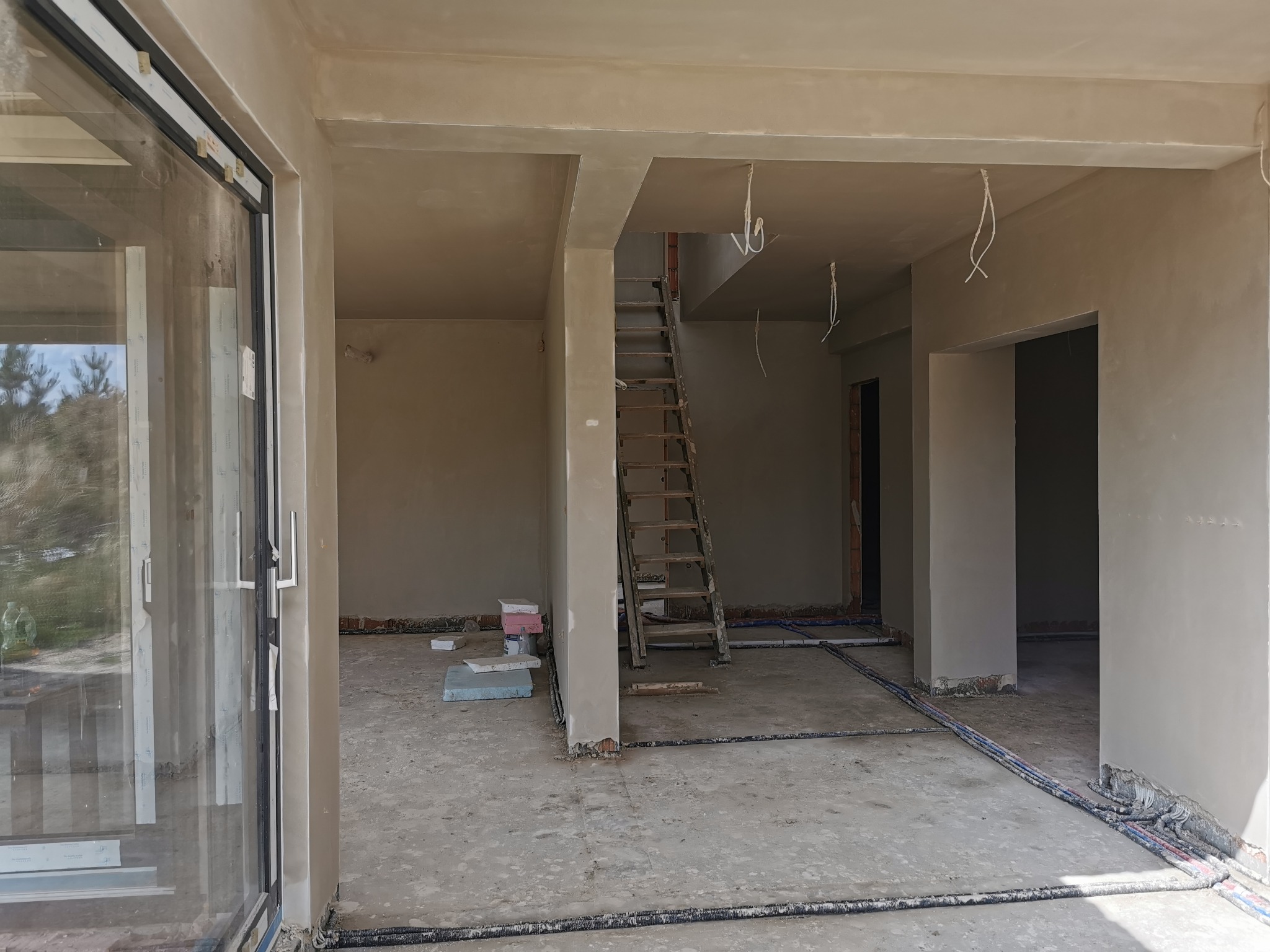 Wnętrze domu w stanie surowym otwartym, ściany pokryte świeżym tynkiem, widoczna drewniana drabina, kable instalacji grzewczej na podłodze i duże okno przesuwne z widokiem na zewnątrz.