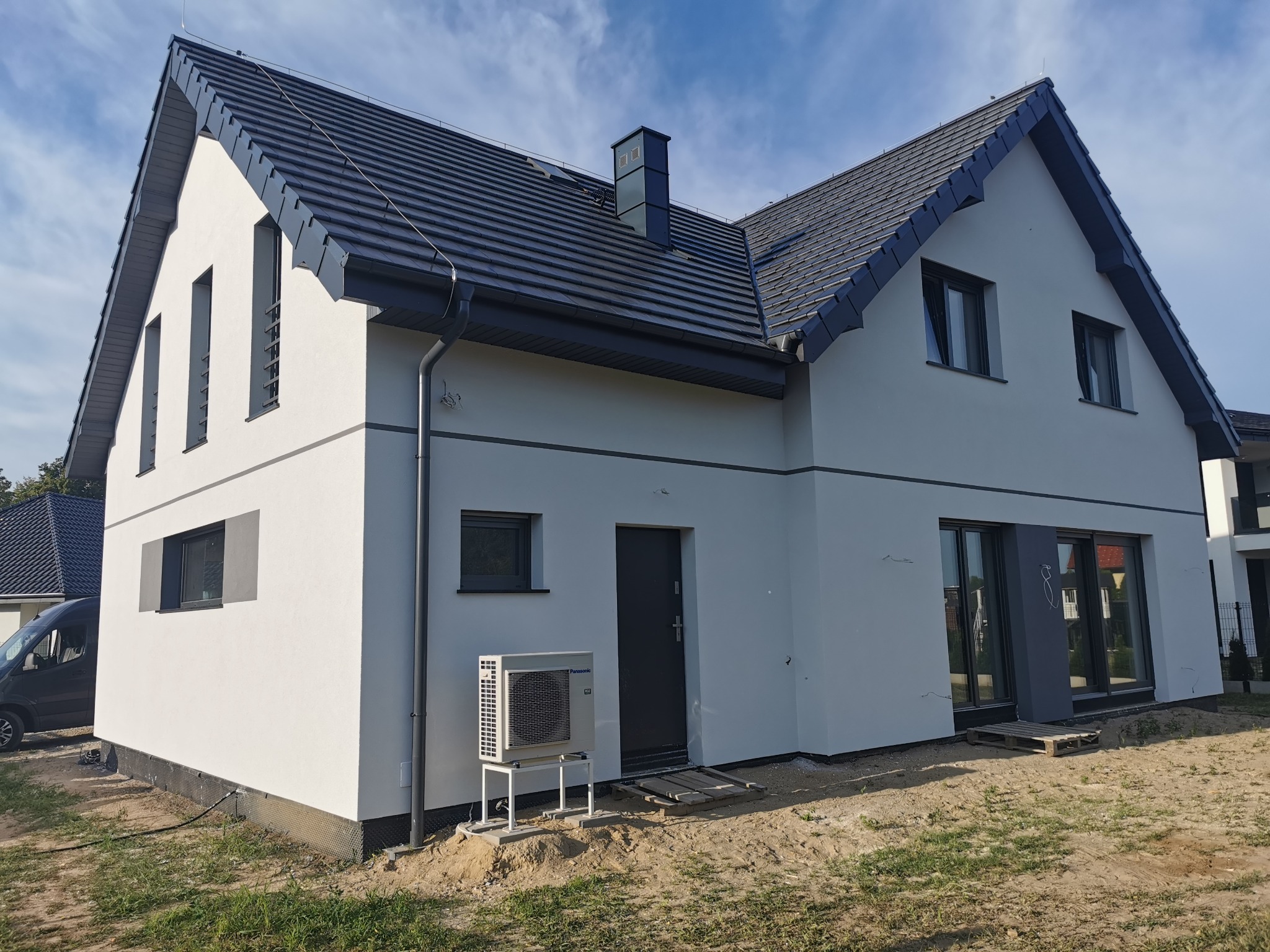 Nowoczesny dom jednorodzinny z białymi ścianami, grafitową stolarką okienną i dachówką, z pompą ciepła Panasonic na zewnątrz i częściowo zagospodarowanym terenem.