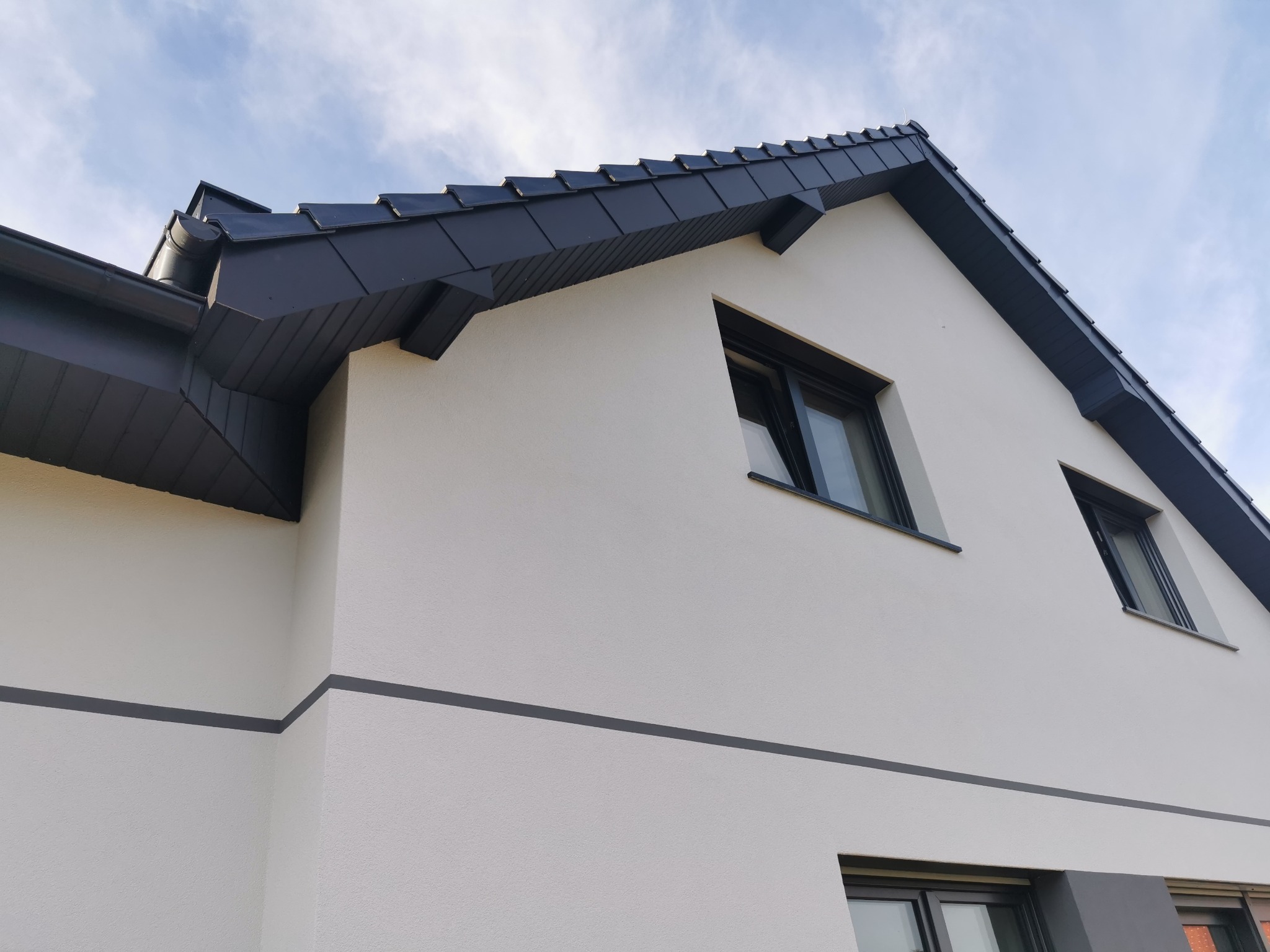 Nowoczesny dom jednorodzinny z białą elewacją, grafitowym dachem i oknami, widok z dołu w słoneczny dzień.