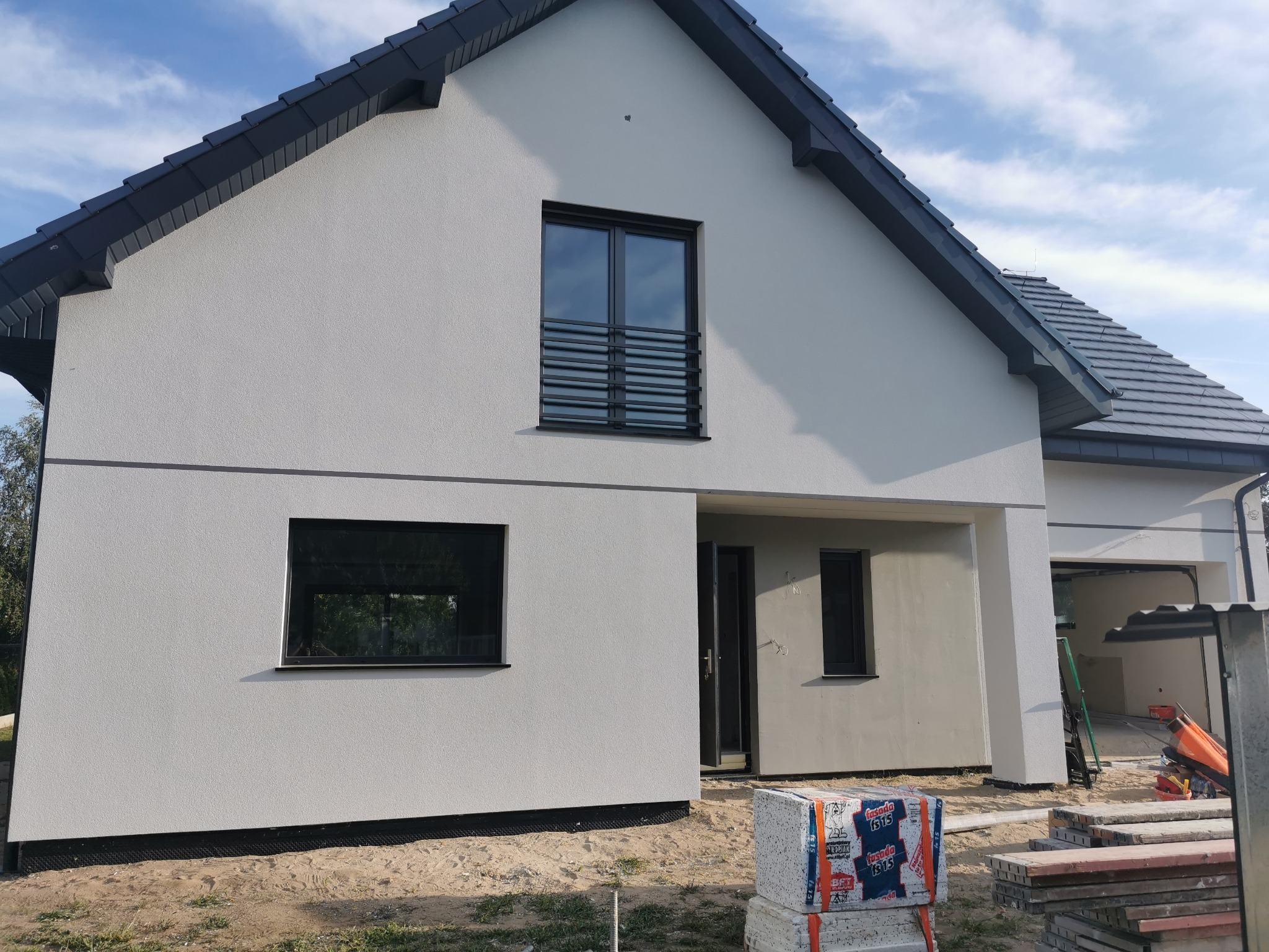 Nowoczesny dom jednorodzinny z jasną elewacją o fakturze baranka, czarną stolarką okienną i grafitową dachówką, widoczne materiały budowlane.