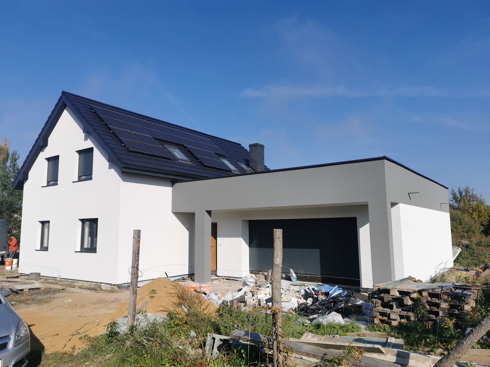 Nowoczesny dom jednorodzinny z białymi ścianami, szarą bryłą garażu i czarną dachówką z panelami solarnymi, widok w słoneczny dzień po zakończonych pracach elewacyjnych, teren wokół jeszcze...