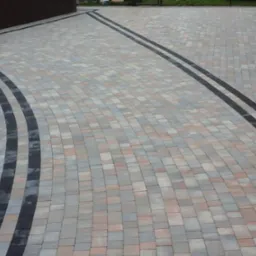 Ułożona kostka brukowa w odcieniach szarości i beżu z czarnymi pasami, tworząca wzór łuku i linii prostych. Widok z góry na ścieżkę.