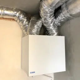 Centrala wentylacyjna z widocznymi, izolowanymi przewodami wentylacyjnymi w surowym wnętrzu z otynkowanymi ścianami.