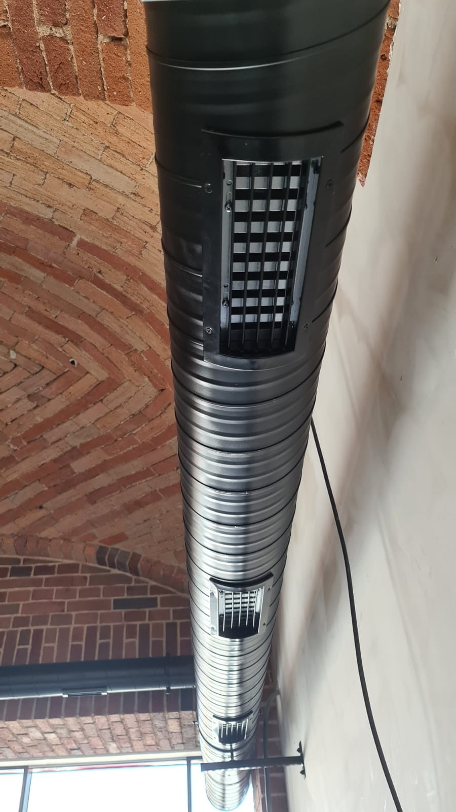 Czarny, spiralny przewód wentylacyjny z kratkami wlotowymi, zamontowany pod ceglanym sklepieniem. Widoczne okno i biała ściana.