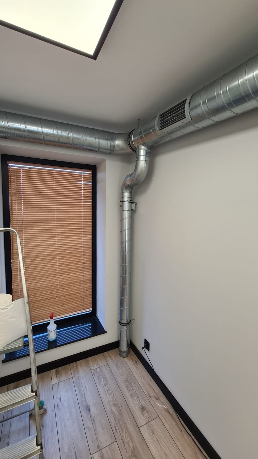 Metalowe rury wentylacyjne zamontowane w rogu pomieszczenia, widoczne okno z żaluzjami i drabina malarska.