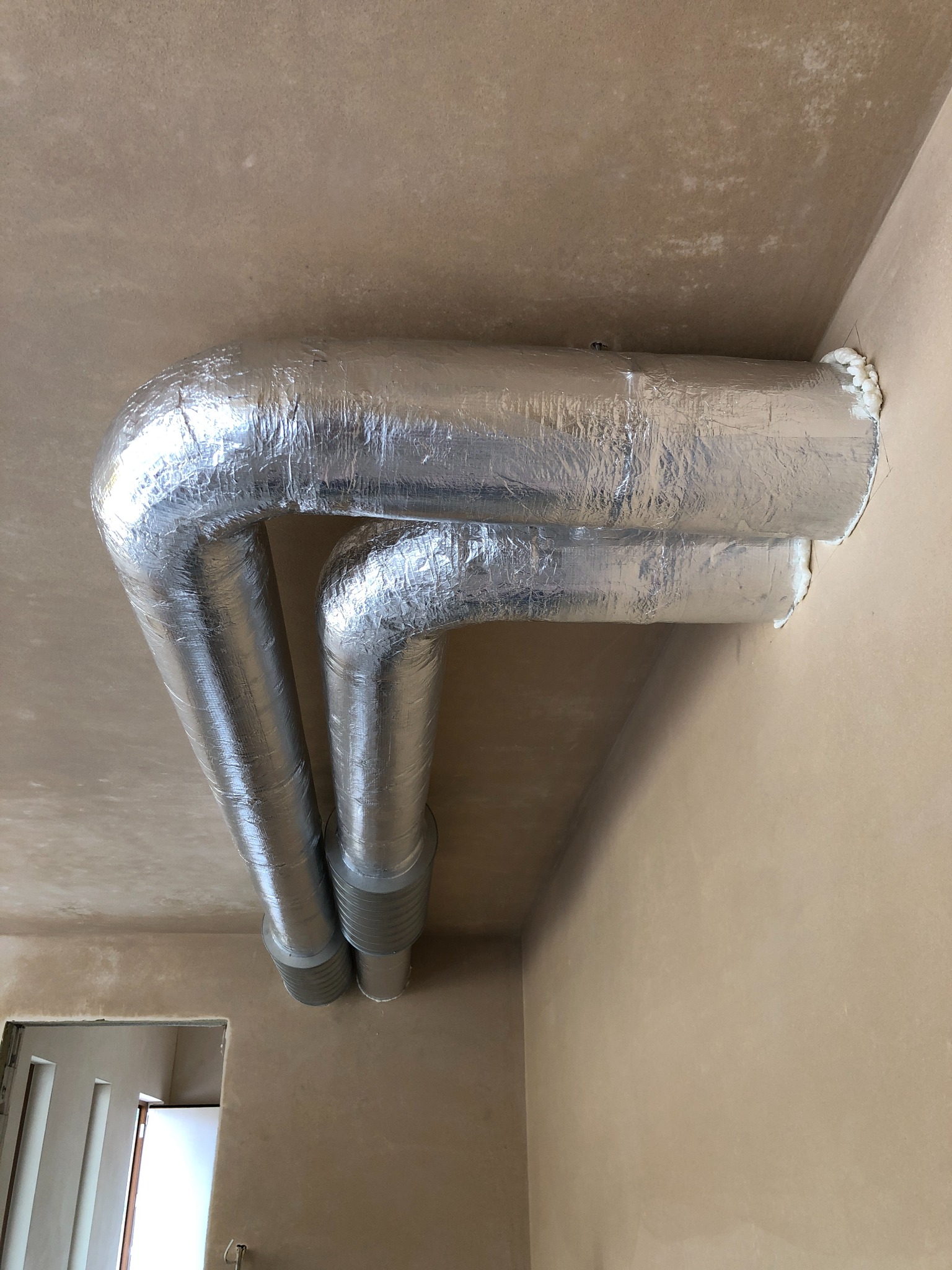 Dwa izolowane przewody wentylacyjne w kształcie litery U, zamontowane pod surowym sufitem, z widoczną pianką uszczelniającą i fragmentem otworu drzwiowego.