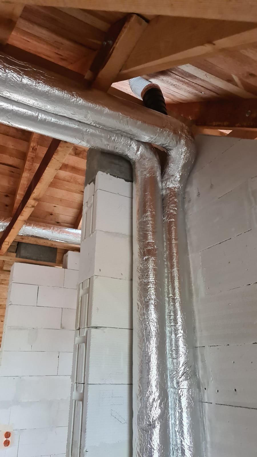 Dwie rury wentylacyjne w srebrnej izolacji prowadzone wzdłuż ściany z białych bloczków, widoczne elementy drewnianej konstrukcji dachu i fragment elastycznego przewodu wentylacyjnego.