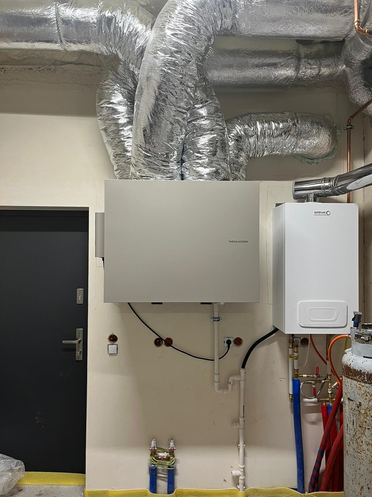 Zamontowana centrala wentylacyjna Thessla Green z widocznymi przewodami wentylacyjnymi spiro i kotłem grzewczym Brötje na ścianie w pomieszczeniu technicznym.