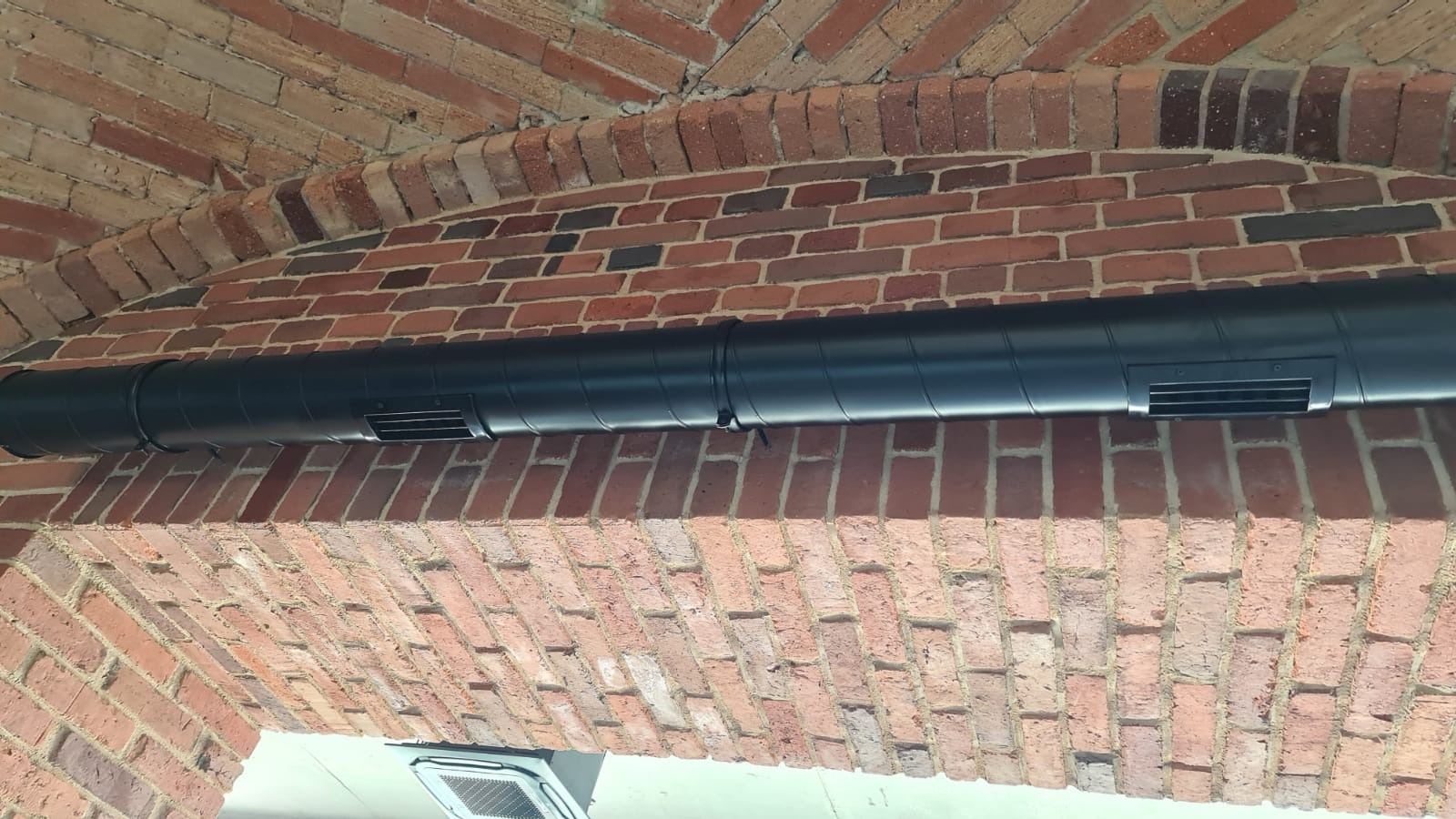 Czarny, okrągły kanał wentylacyjny zamontowany pod ceglanym łukiem. Widoczne kratki wentylacyjne na kanale.