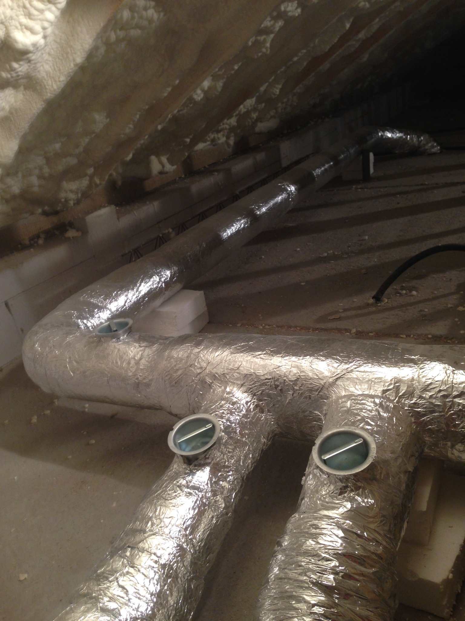 Izolowane kanały wentylacyjne z metalowymi kratkami w przestrzeni poddasza, widoczna pianka izolacyjna na ścianach.