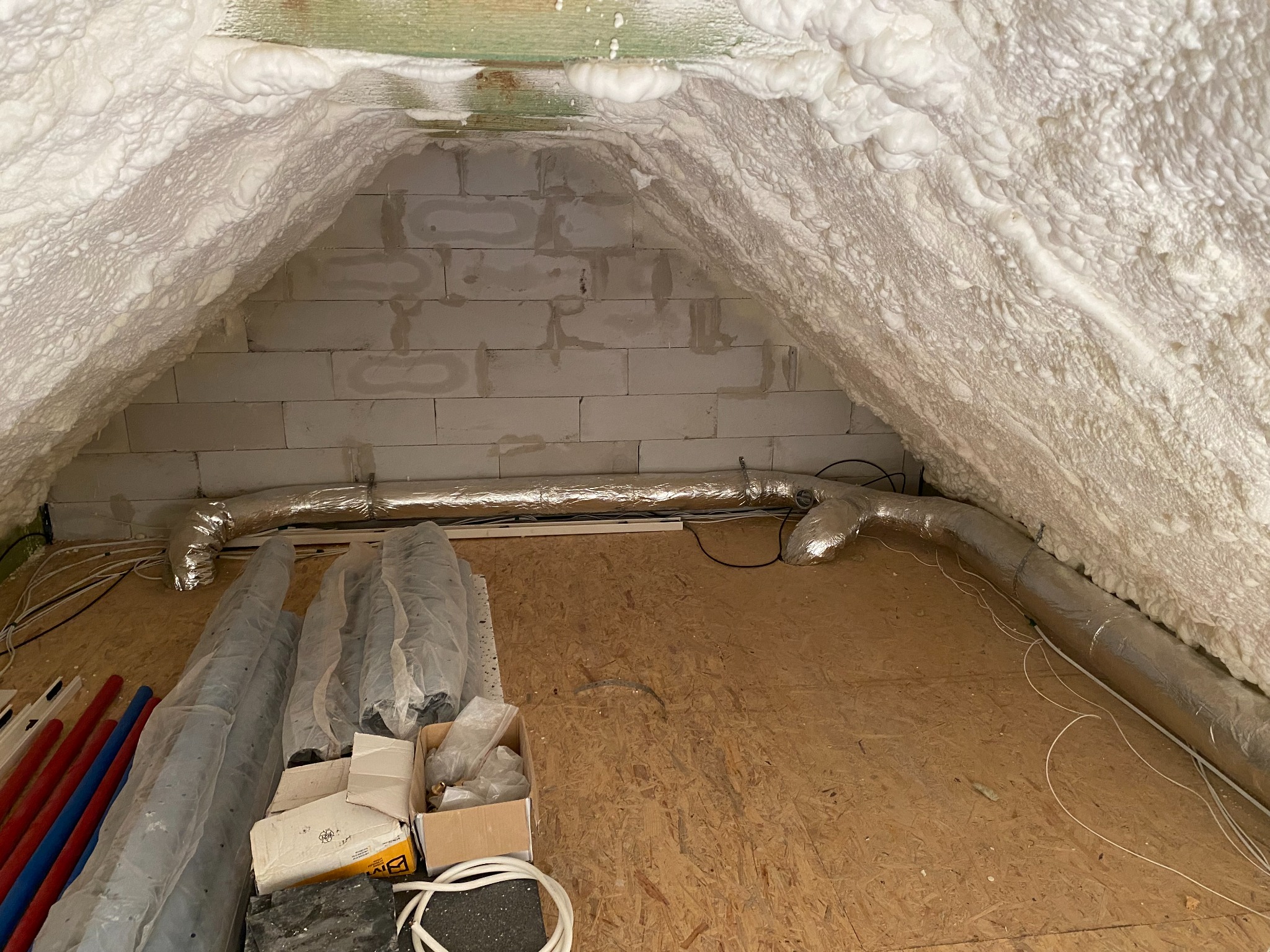 Poddasze ocieplone pianą poliuretanową, widoczne fragmenty instalacji wentylacyjnej z rurami w izolacji oraz materiały budowlane na podłodze z płyty OSB.