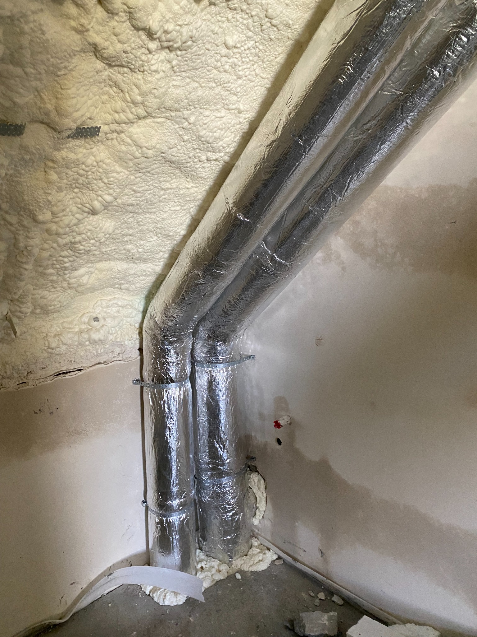 Dwie aluminiowe rury wentylacyjne przymocowane do ściany i sufitu pokrytego pianką izolacyjną w surowym wnętrzu.