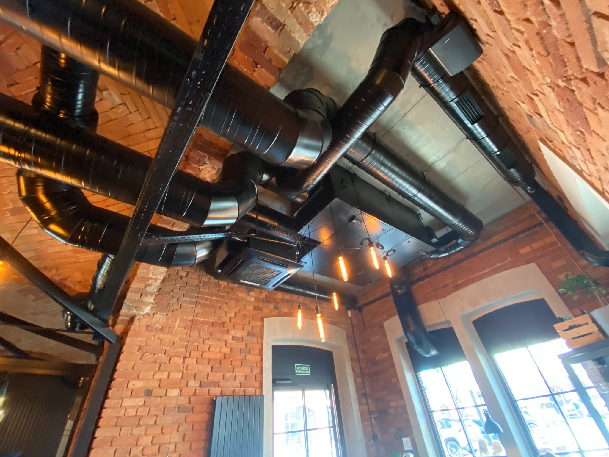 Widok z dołu na czarne rury wentylacyjne zamontowane pod sufitem w pomieszczeniu z ceglanymi ścianami i wiszącymi lampami