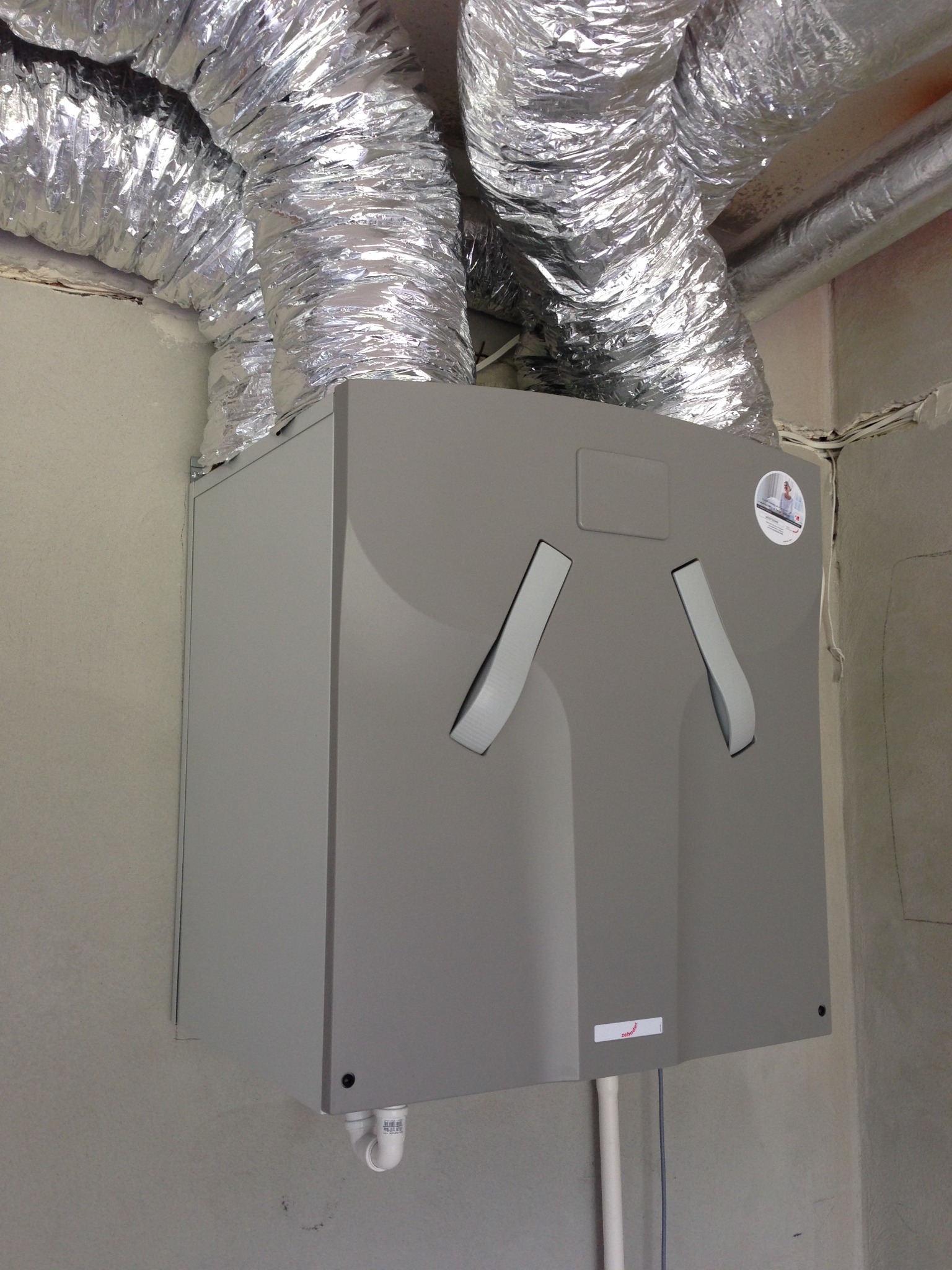 Szara centrala wentylacyjna z podłączonymi, elastycznymi przewodami aluminiowymi w surowym wnętrzu. Widoczne otwory wentylacyjne i naklejka producenta.