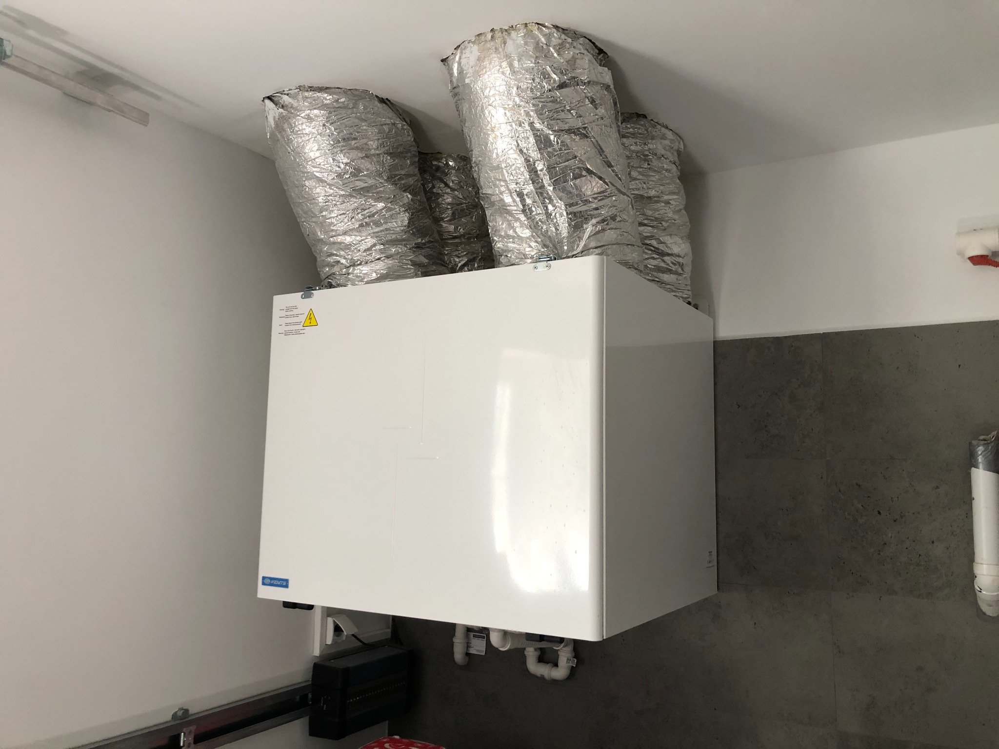 Centralna jednostka wentylacyjna z czterema przewodami wentylacyjnymi w izolacji aluminiowej, zamontowana w pomieszczeniu z szarą ścianą.