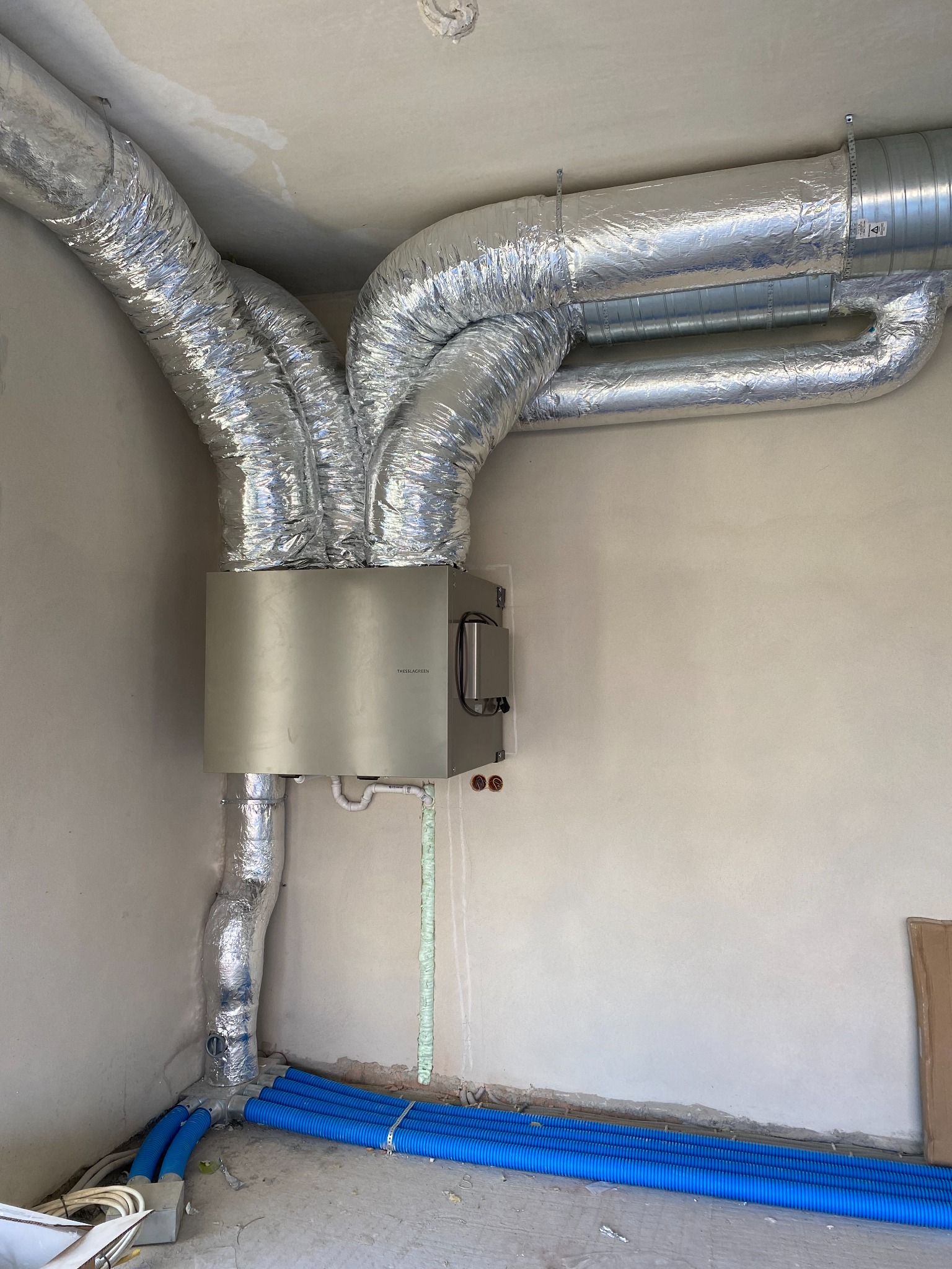 Szare urządzenie rekuperacyjne marki Thessla Green z podłączonymi, zaizolowanymi przewodami wentylacyjnymi w surowym wnętrzu, z niebieskimi rurami karbowanymi na podłodze.