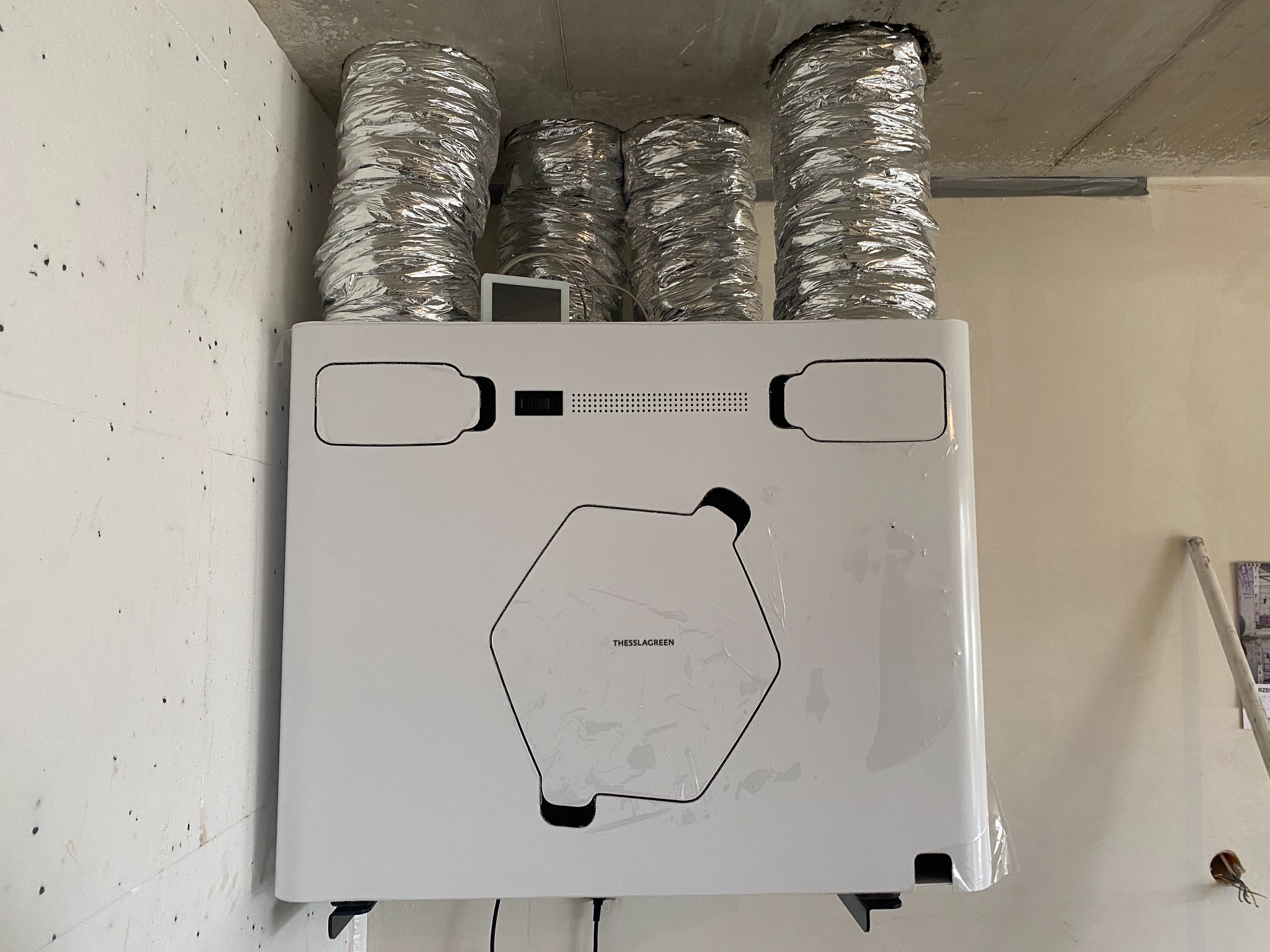 Biała centrala wentylacyjna Thessla Green z czterema podłączonymi, elastycznymi przewodami wentylacyjnymi w aluminiowej osłonie, zamontowana na ścianie w surowym wnętrzu.