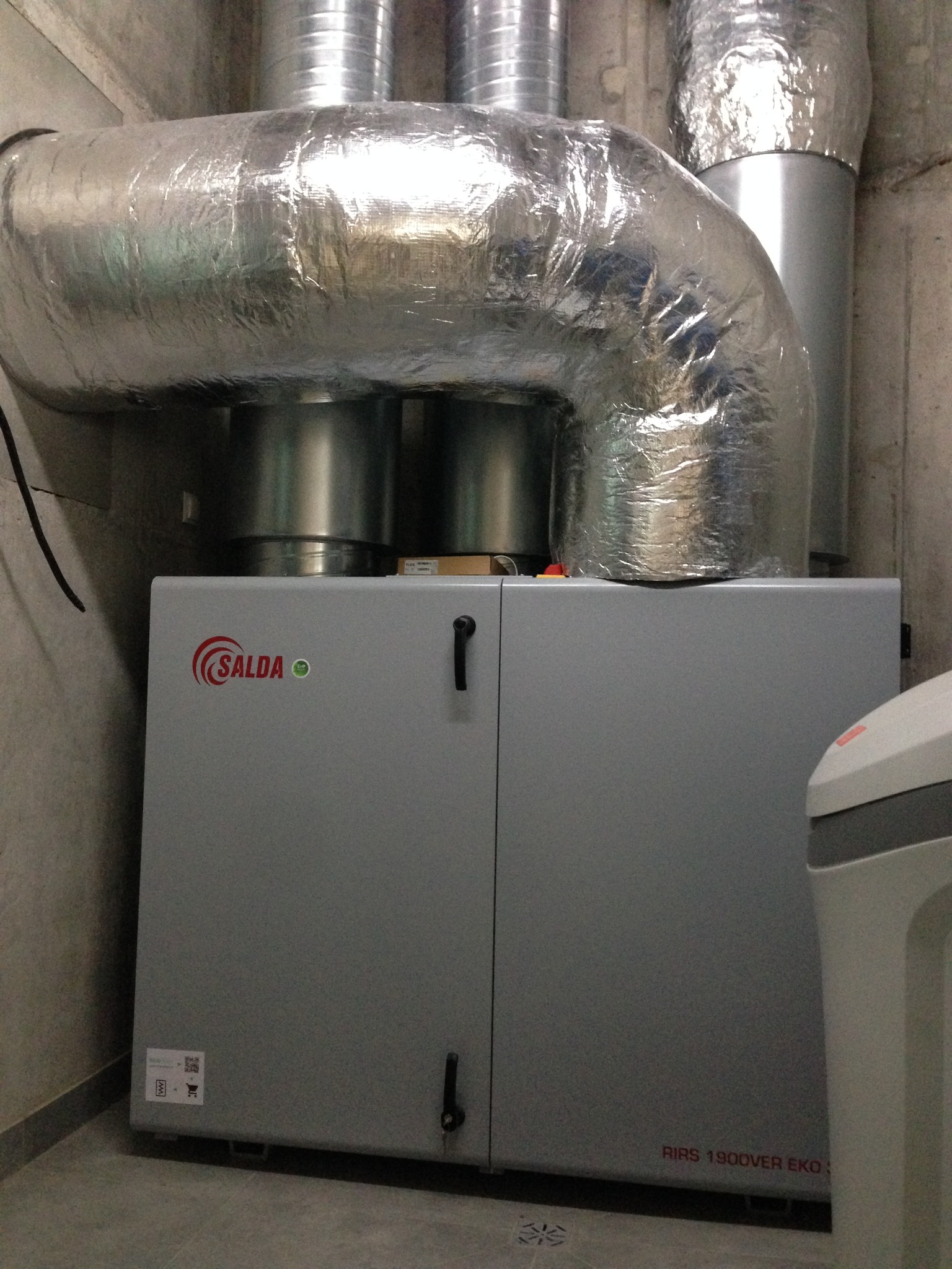 Szara centrala wentylacyjna Salda RIRS 1900VER EKO z izolowanymi przewodami wentylacyjnymi w pomieszczeniu technicznym.
