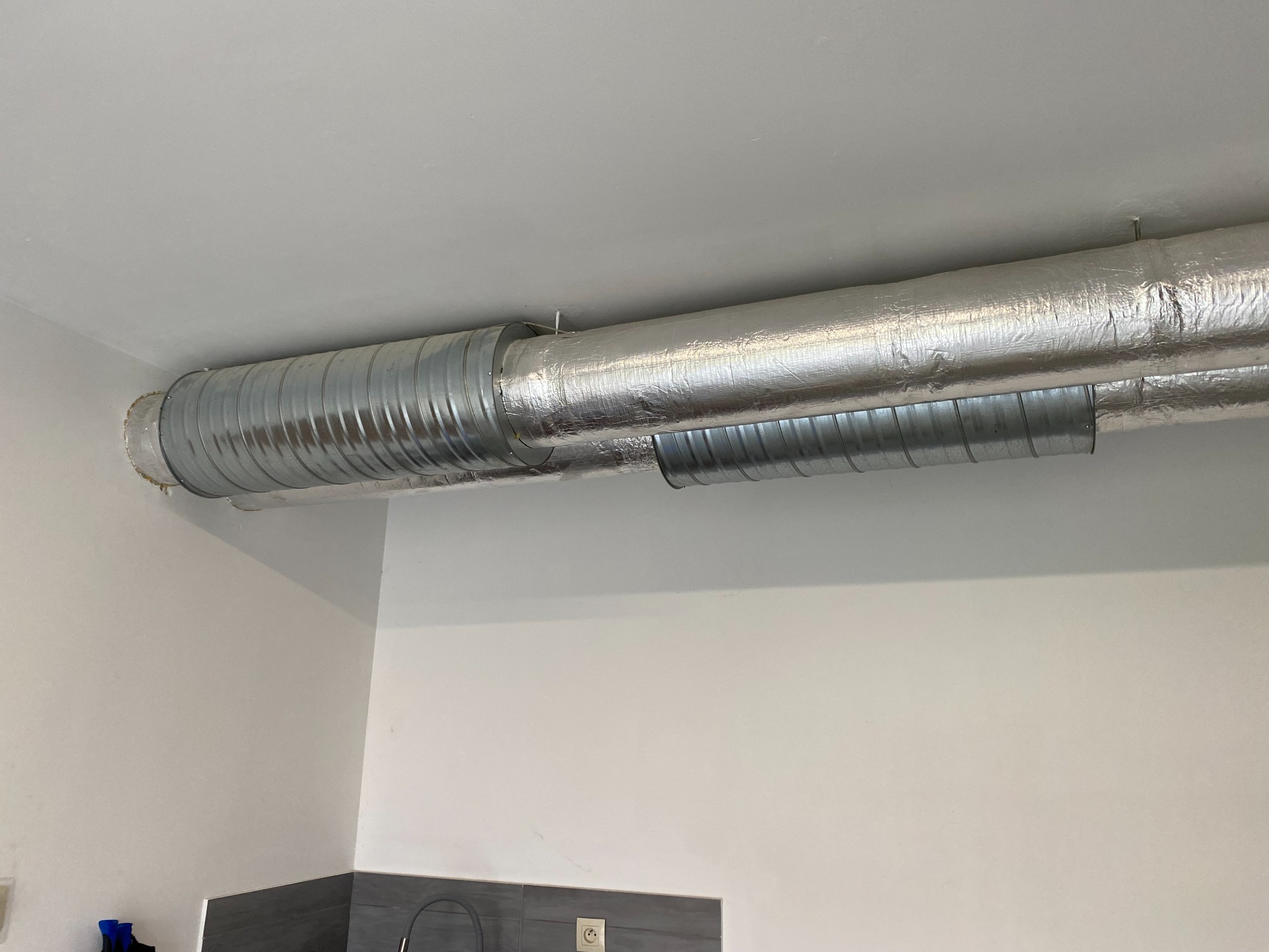 Metalowa rura wentylacyjna przymocowana do sufitu w pomieszczeniu z białymi ścianami i szarymi elementami wyposażenia.
