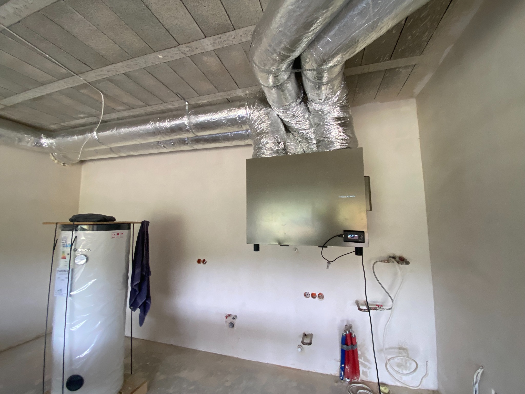 Wnętrze w trakcie prac instalacyjnych: srebrne rury wentylacyjne pod betonowym sufitem, centrala wentylacyjna na ścianie, bojler z niebieskim ręcznikiem obok i wystające rury instalacji wodnej.