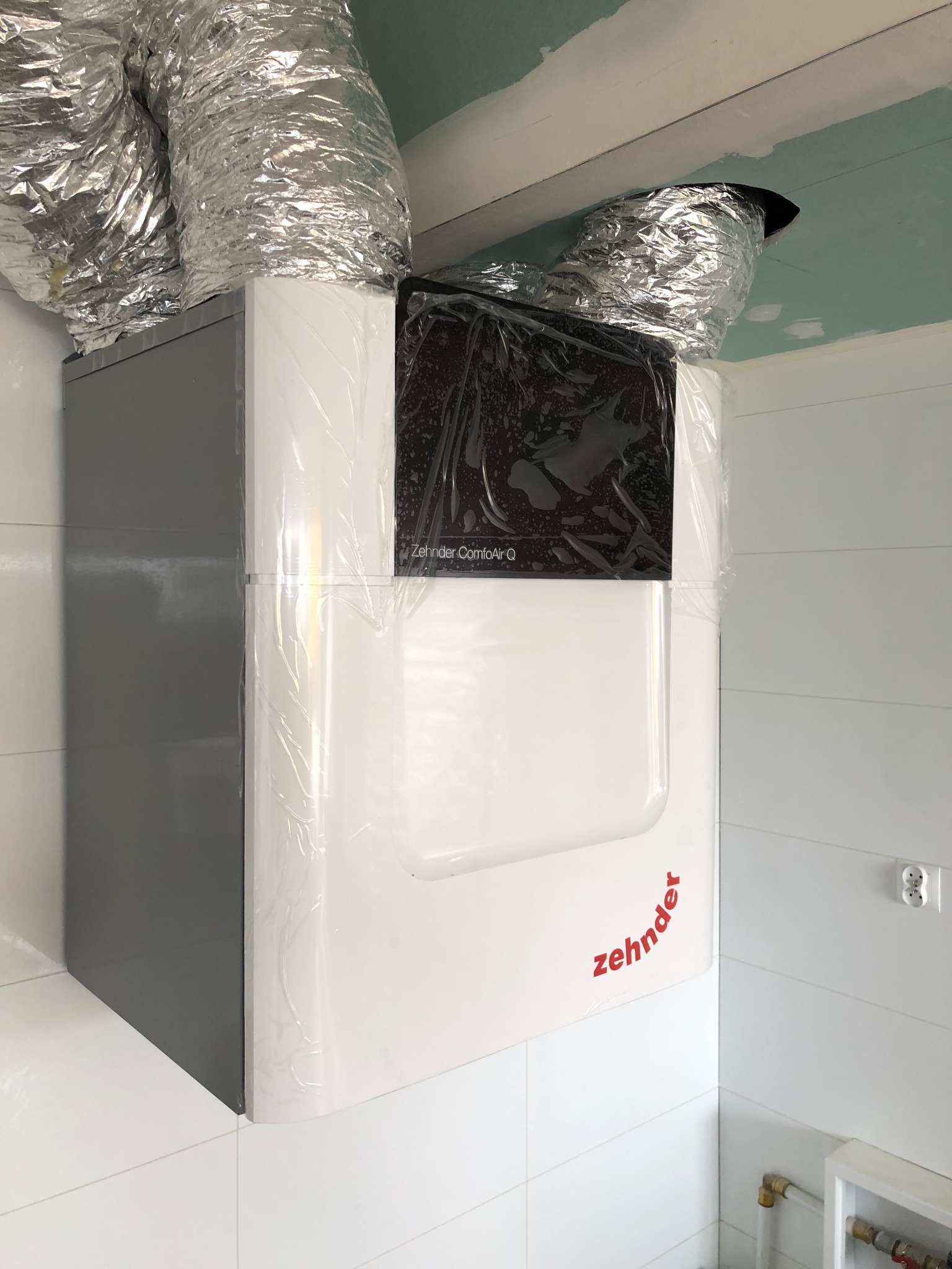 Centrala wentylacyjna z rekuperacją marki Zehnder ComfoAir Q, z podłączonymi przewodami wentylacyjnymi w folii ochronnej, zamontowana w pomieszczeniu z białymi płytkami.