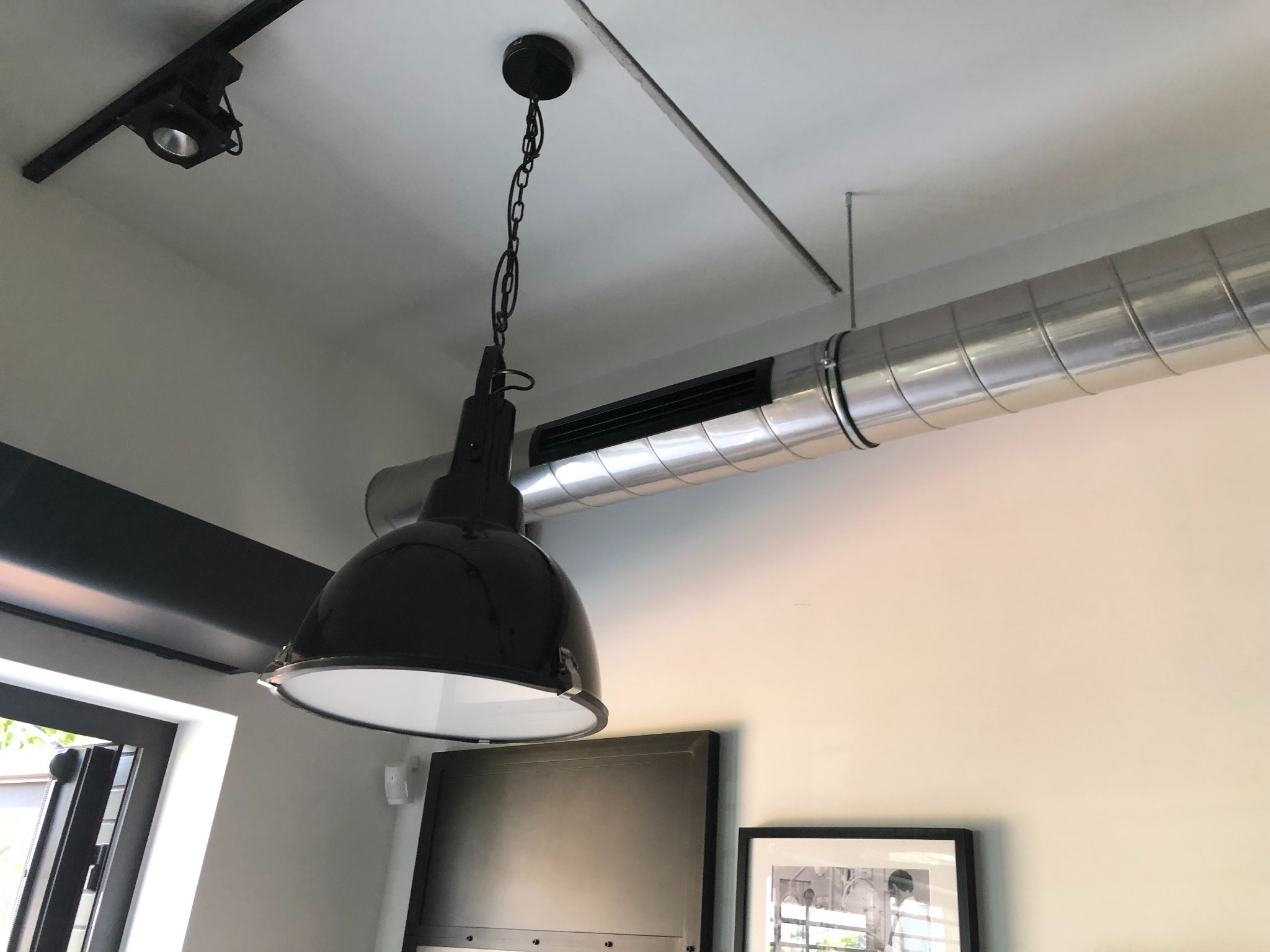 Czarny, wiszący reflektor przemysłowy na łańcuchu, widoczny na tle rury wentylacyjnej, szyny oświetleniowej i fragmentu okna.