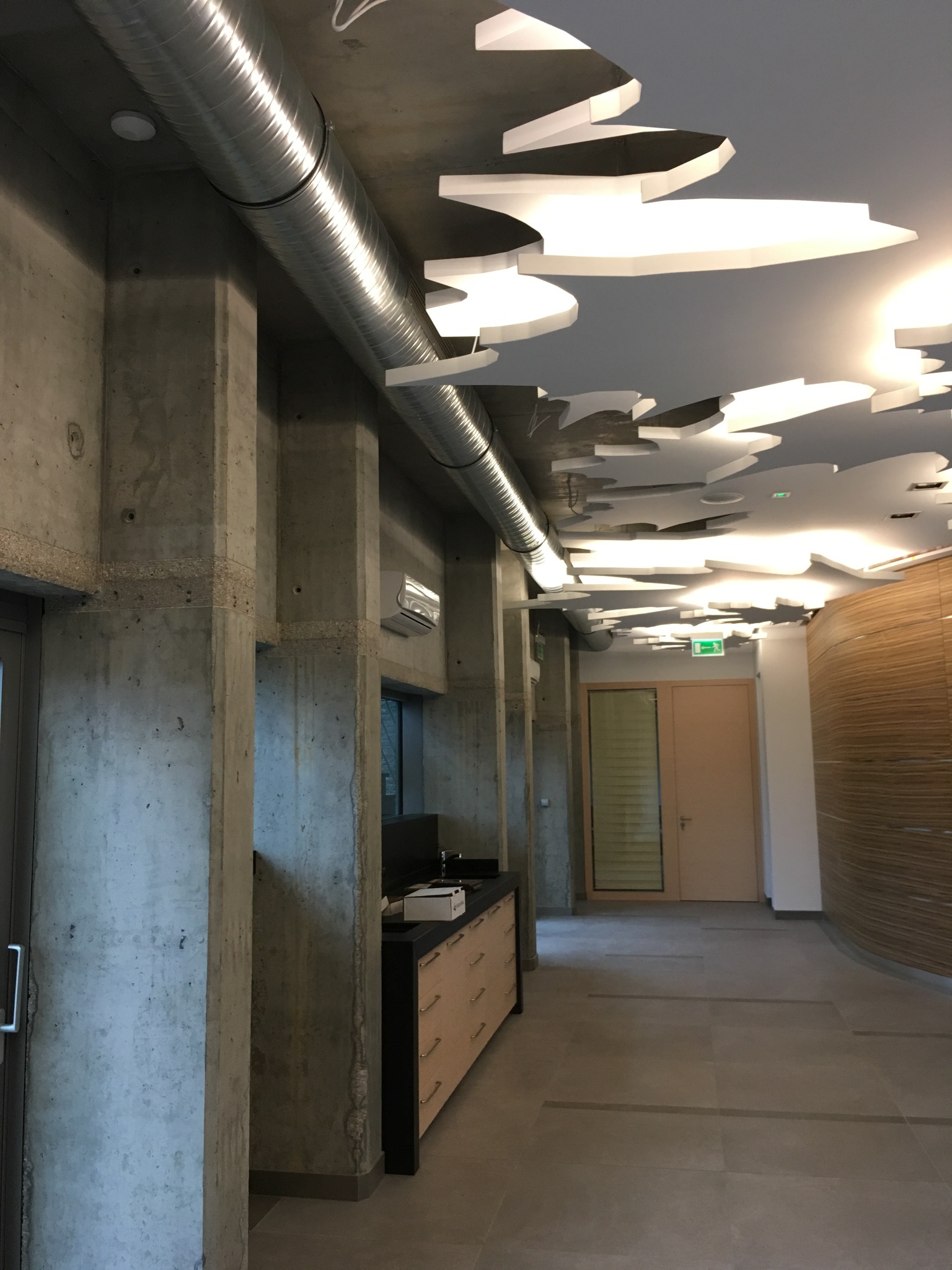 Wnętrze biurowe z widoczną instalacją wentylacyjną pod sufitem, betonowe kolumny, dekoracyjny sufit podwieszany z oświetleniem, szafka z szufladami przy ścianie.