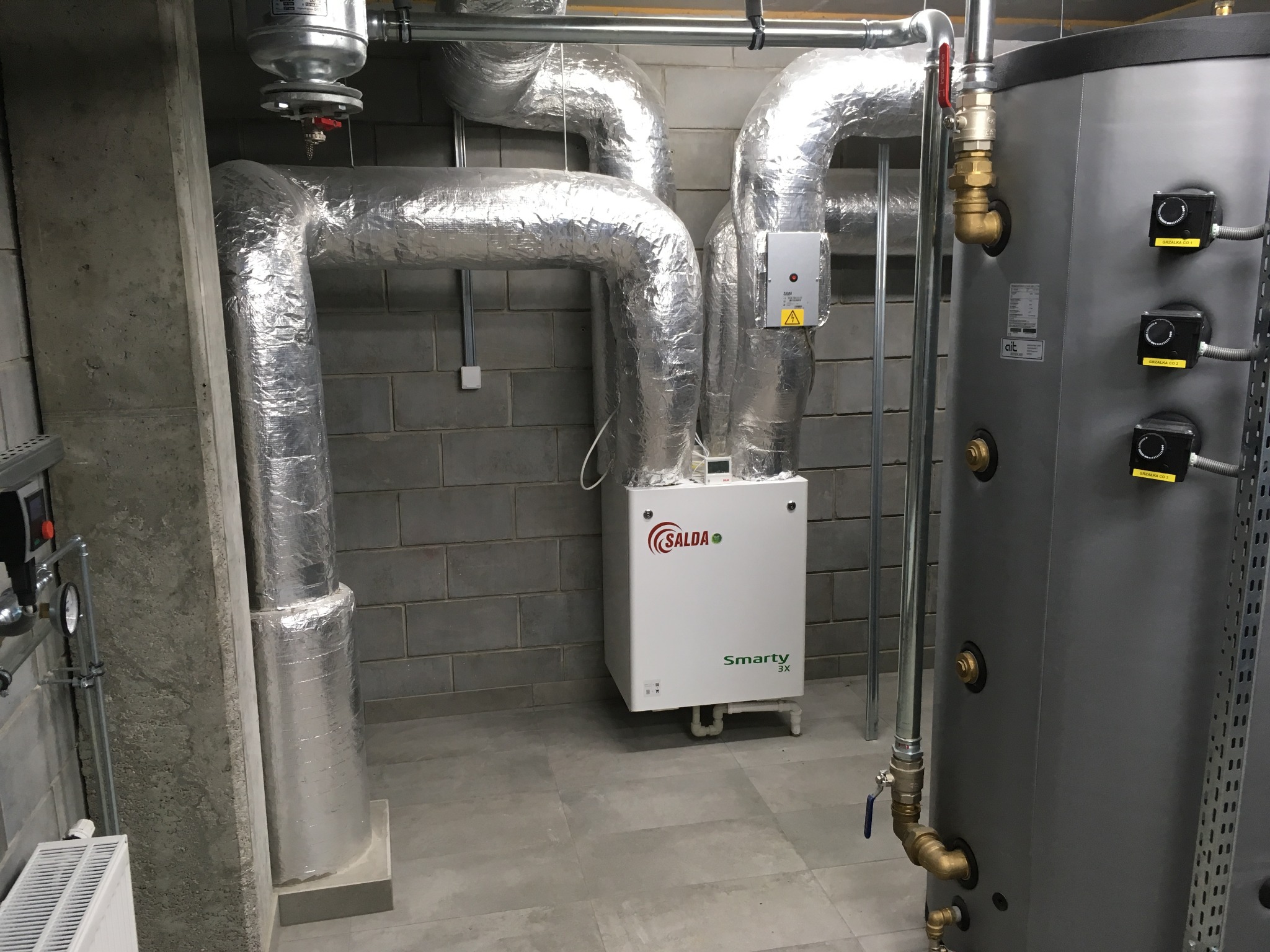 Instalacja systemu wentylacji z izolowanymi przewodami, jednostką centralną SALDA Smarty 3X i zasobnikiem ciepłej wody w szarym pomieszczeniu technicznym.