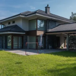 Dwukondygnacyjny dom murowany z grafitową dachówką i kamienną elewacją, widok z ogrodu z bramką do piłki nożnej i zadaszonym tarasem z meblami ogrodowymi.