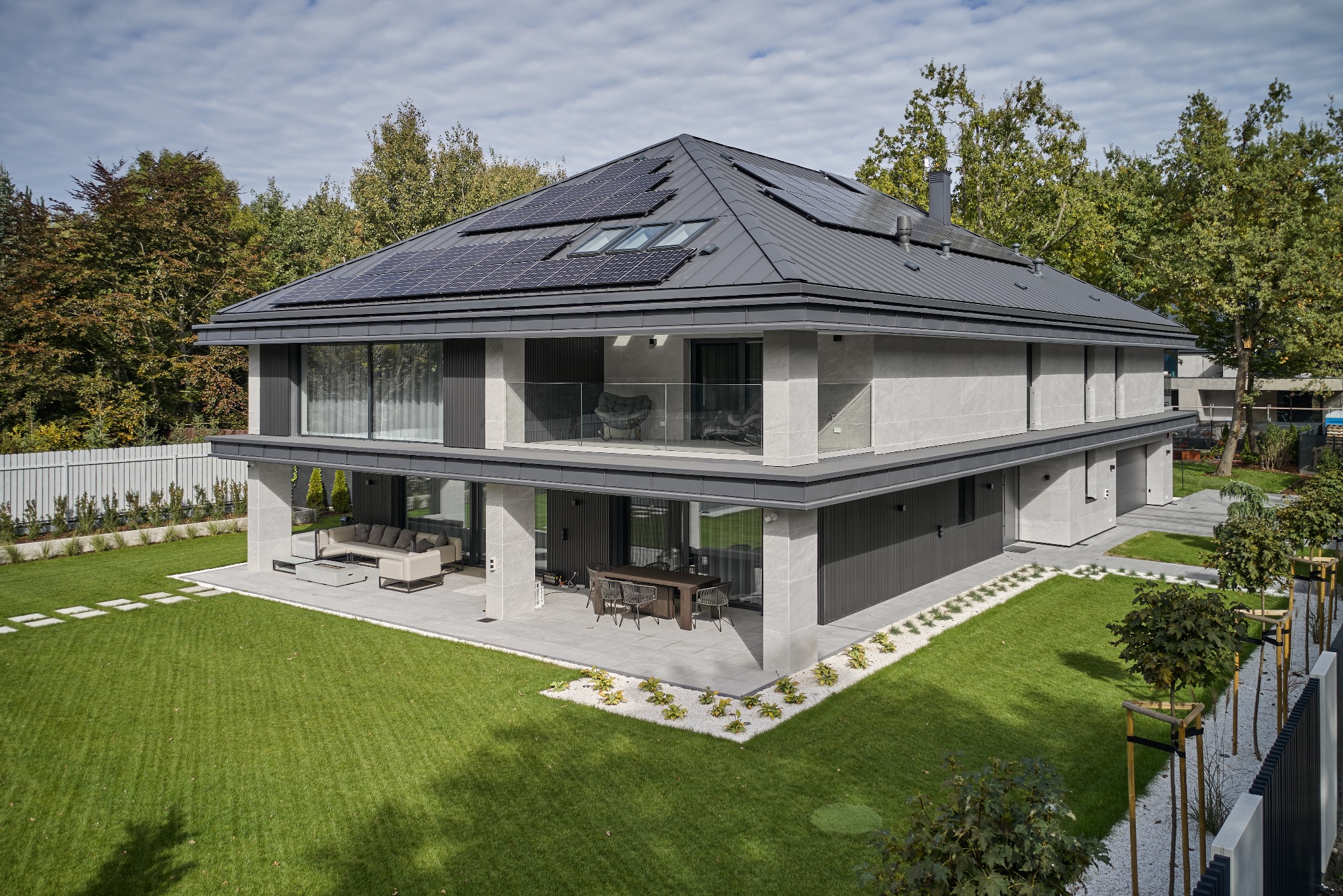 Dwukondygnacyjny dom murowany z płaskim dachem pokrytym panelami słonecznymi, z dużymi przeszkleniami i tarasem z meblami ogrodowymi, otoczony zadbanym trawnikiem i drzewami.