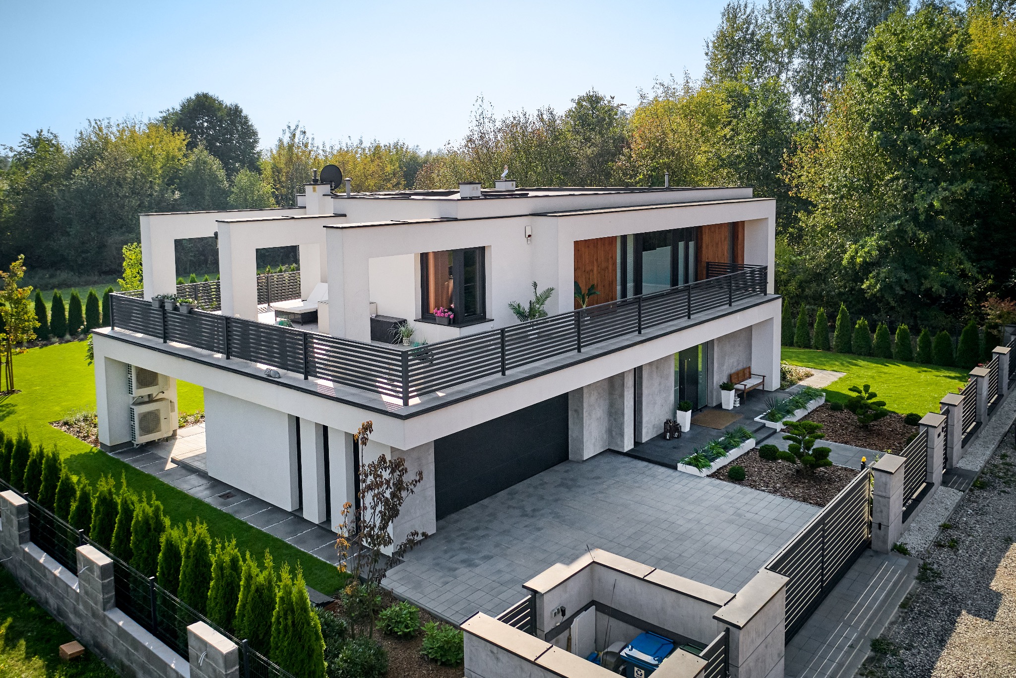 Nowoczesny, dwukondygnacyjny dom z płaskim dachem i dużym tarasem, widok z góry na zadbany ogród i podjazd z kostki brukowej.