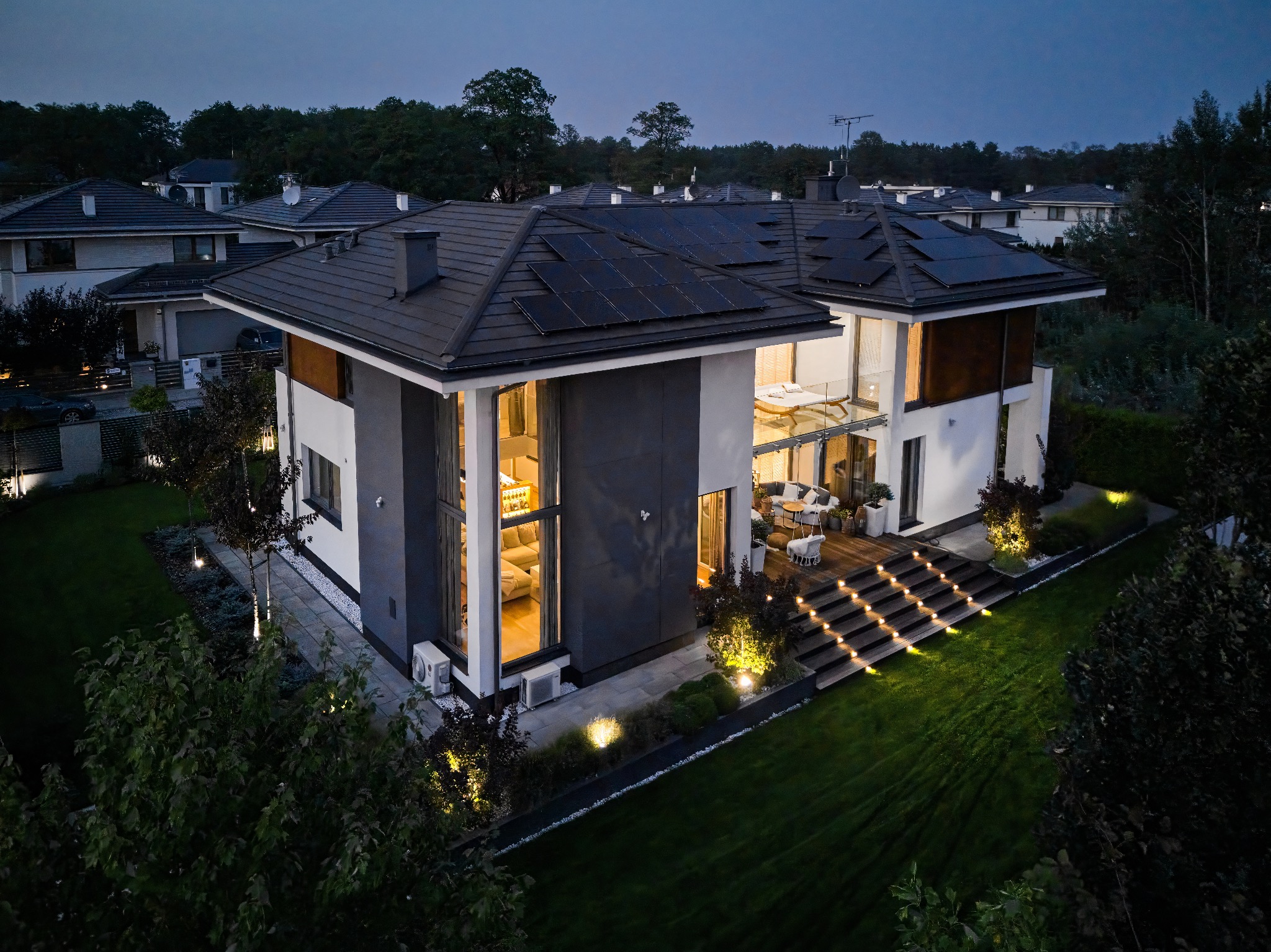 Nowoczesny dom jednorodzinny z panelami słonecznymi na dachu, oświetlony zewnętrznymi lampami, widok z lotu ptaka o zmierzchu.