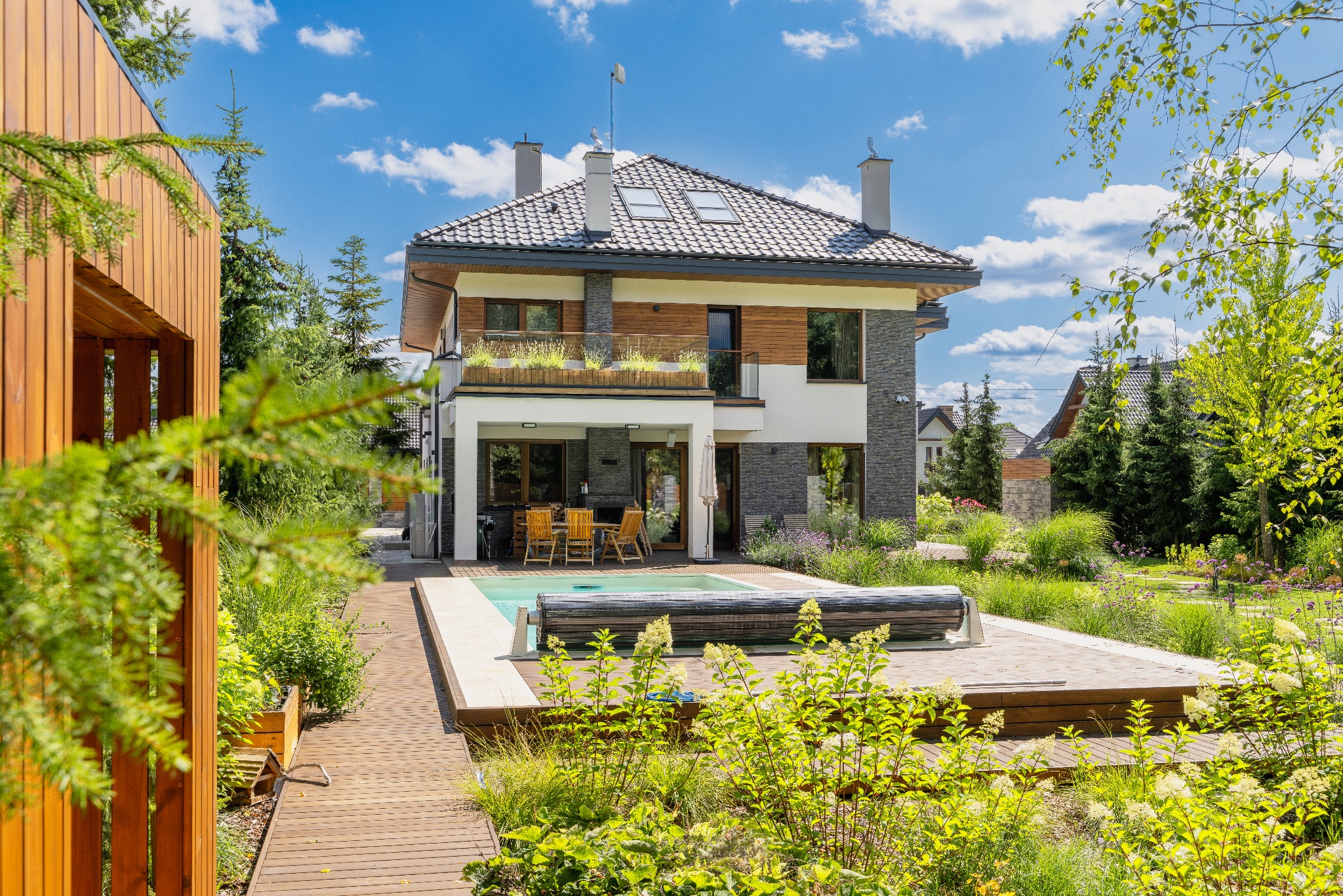 Dwukondygnacyjny dom z szarą dachówką, wykończony elementami z drewna i kamienia, z tarasem, basenem i zadbanym ogrodem, widziany w słoneczny dzień.