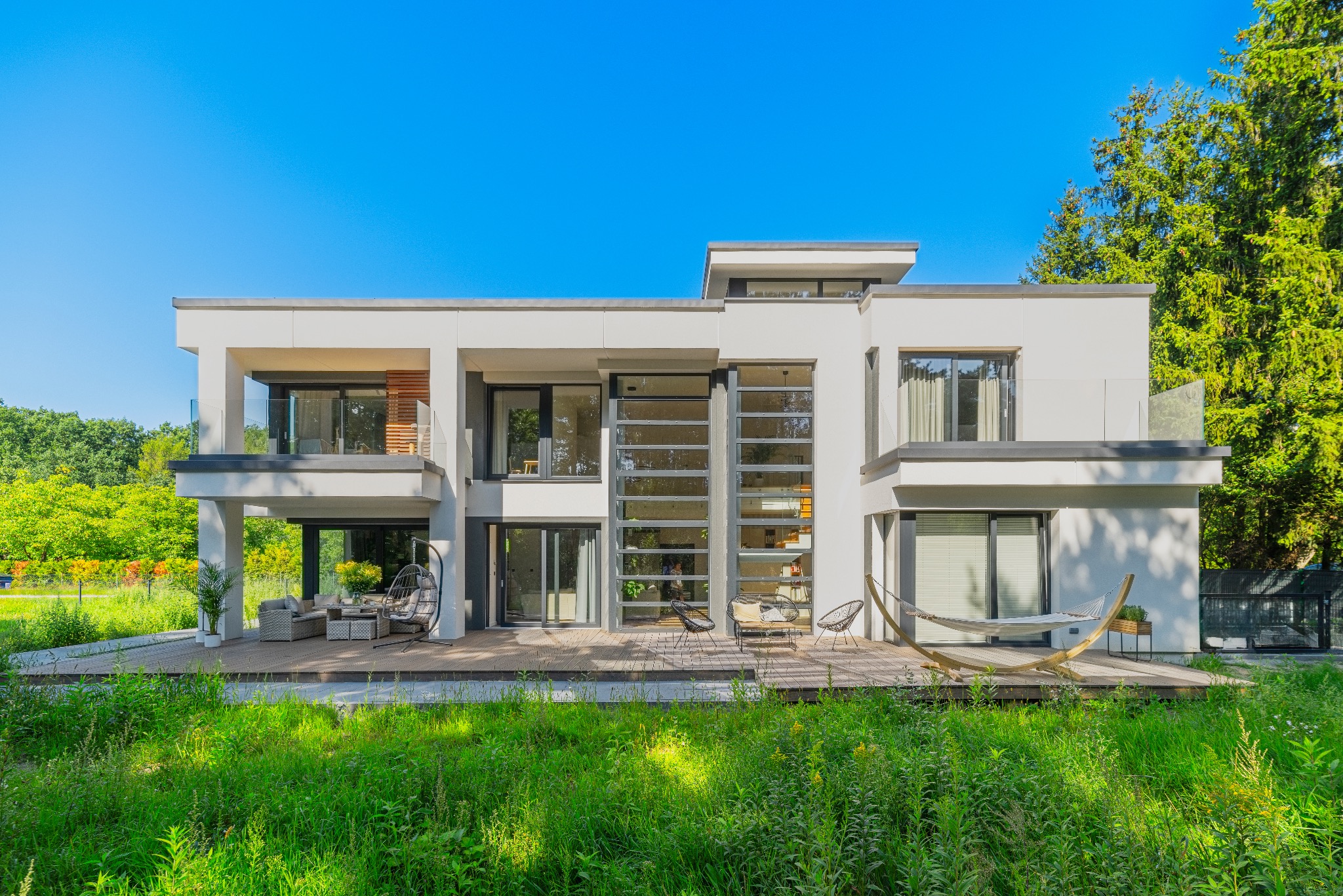 Nowoczesny, dwukondygnacyjny dom murowany z płaskim dachem, dużymi oknami i tarasem z meblami wypoczynkowymi oraz hamakiem, otoczony zielenią.