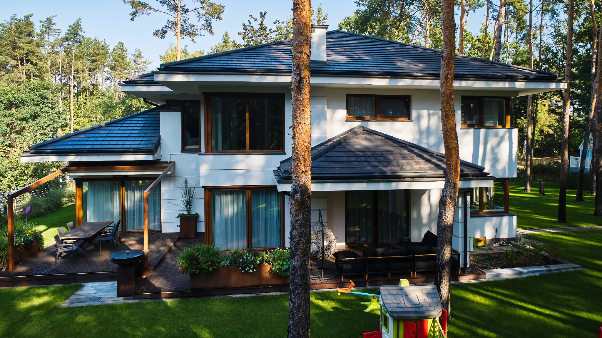 Nowoczesny, dwukondygnacyjny dom z białymi ścianami i ciemną dachówką, otoczony zielonym trawnikiem i drzewami. Na tarasie widoczny stół z krzesłami i grill, a na drugim tarasie czarna sofa...