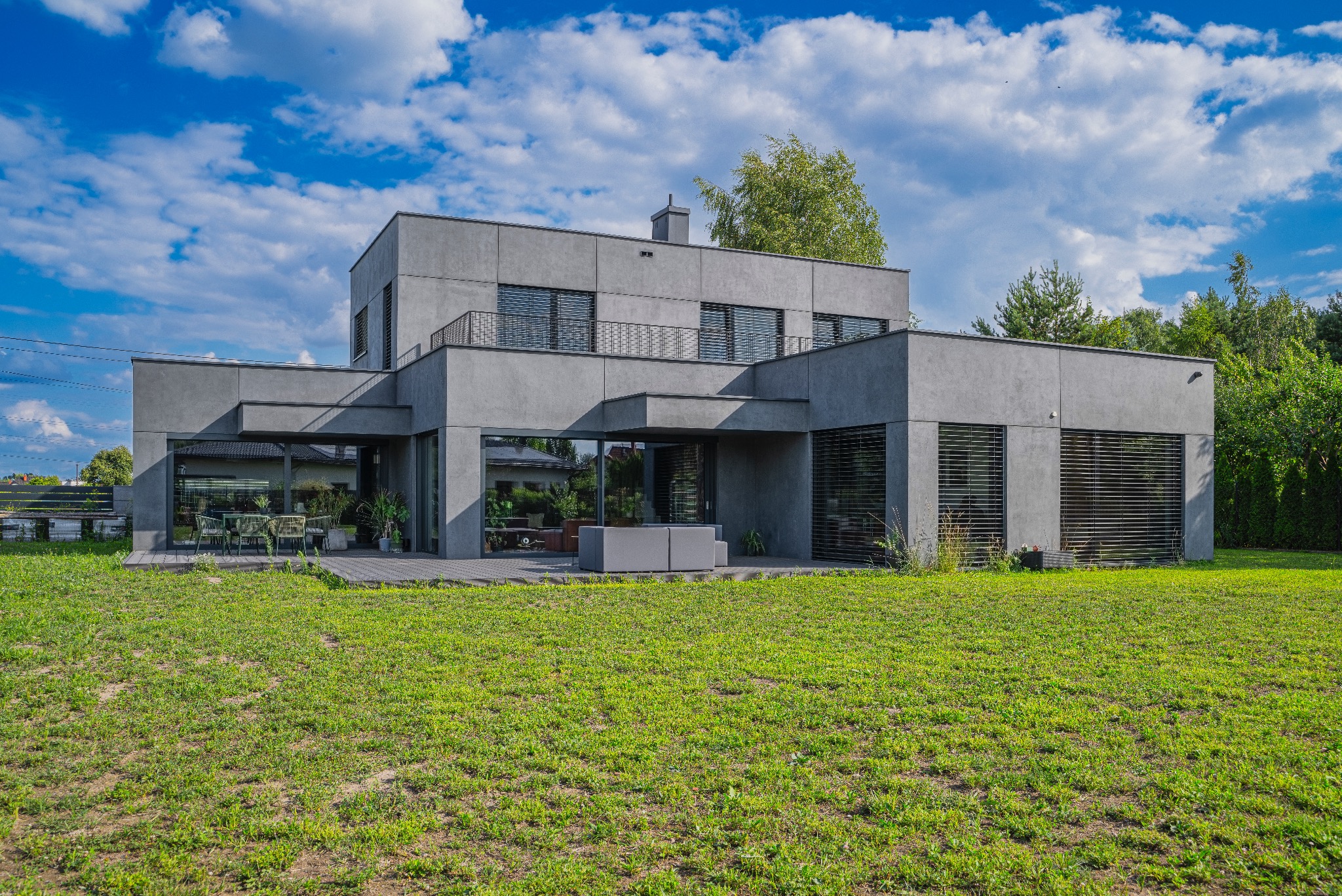 Nowoczesny, dwukondygnacyjny dom murowany w odcieniach szarości z płaskim dachem, widoczny z perspektywy ogrodu, z wyraźnymi dużymi oknami i elementami wykończenia w stylu minimalistycznym, na tle...