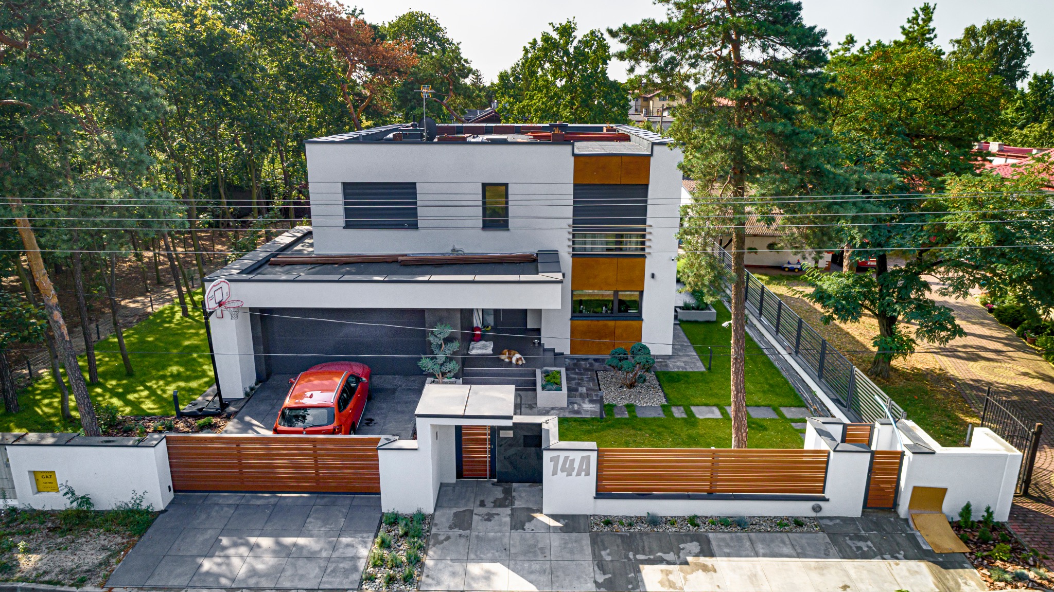 Nowoczesny dom jednorodzinny z płaskim dachem, garażem, elementami dekoracyjnymi z drewna i numerem 14A na ogrodzeniu, widziany z góry, z czerwonym samochodem zaparkowanym w garażu i psem leżącym...