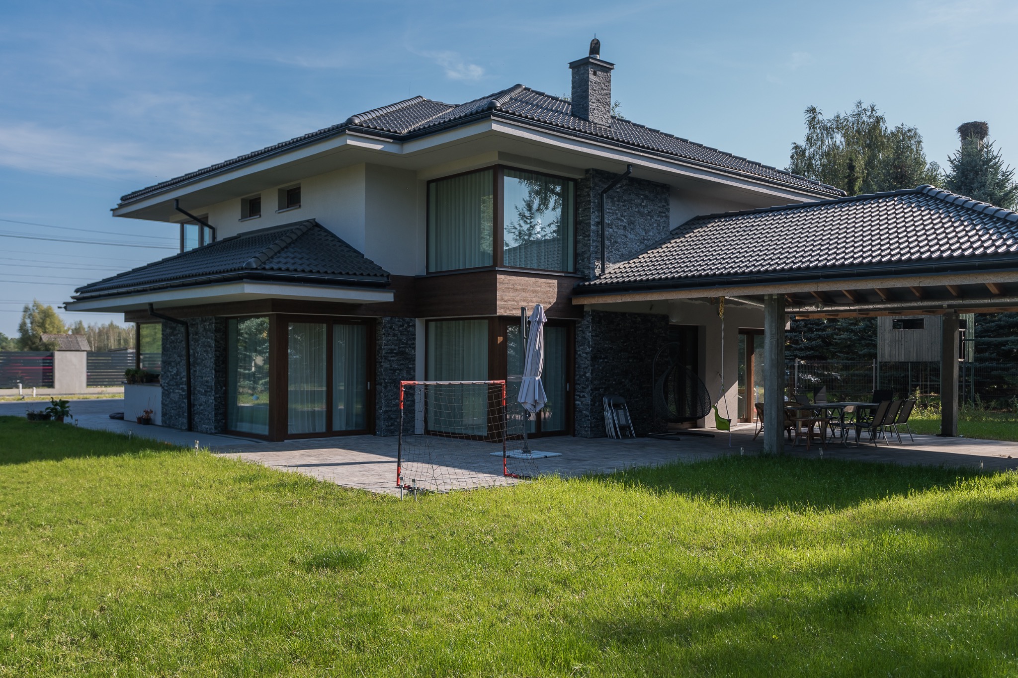 Dwukondygnacyjny dom murowany z grafitową dachówką i kamienną elewacją, widok z ogrodu z bramką do piłki nożnej i zadaszonym tarasem z meblami ogrodowymi.